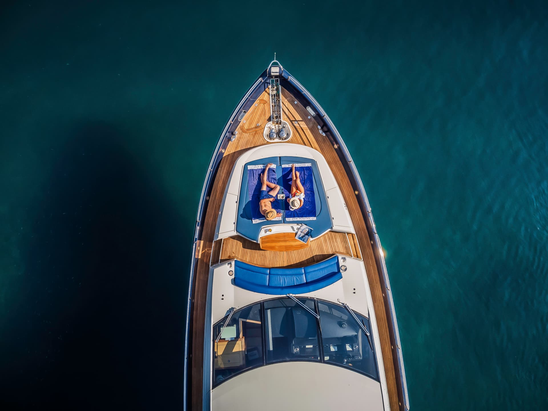 Yacht with two people sunbathing on the bow over deep turquoise coastal waters.