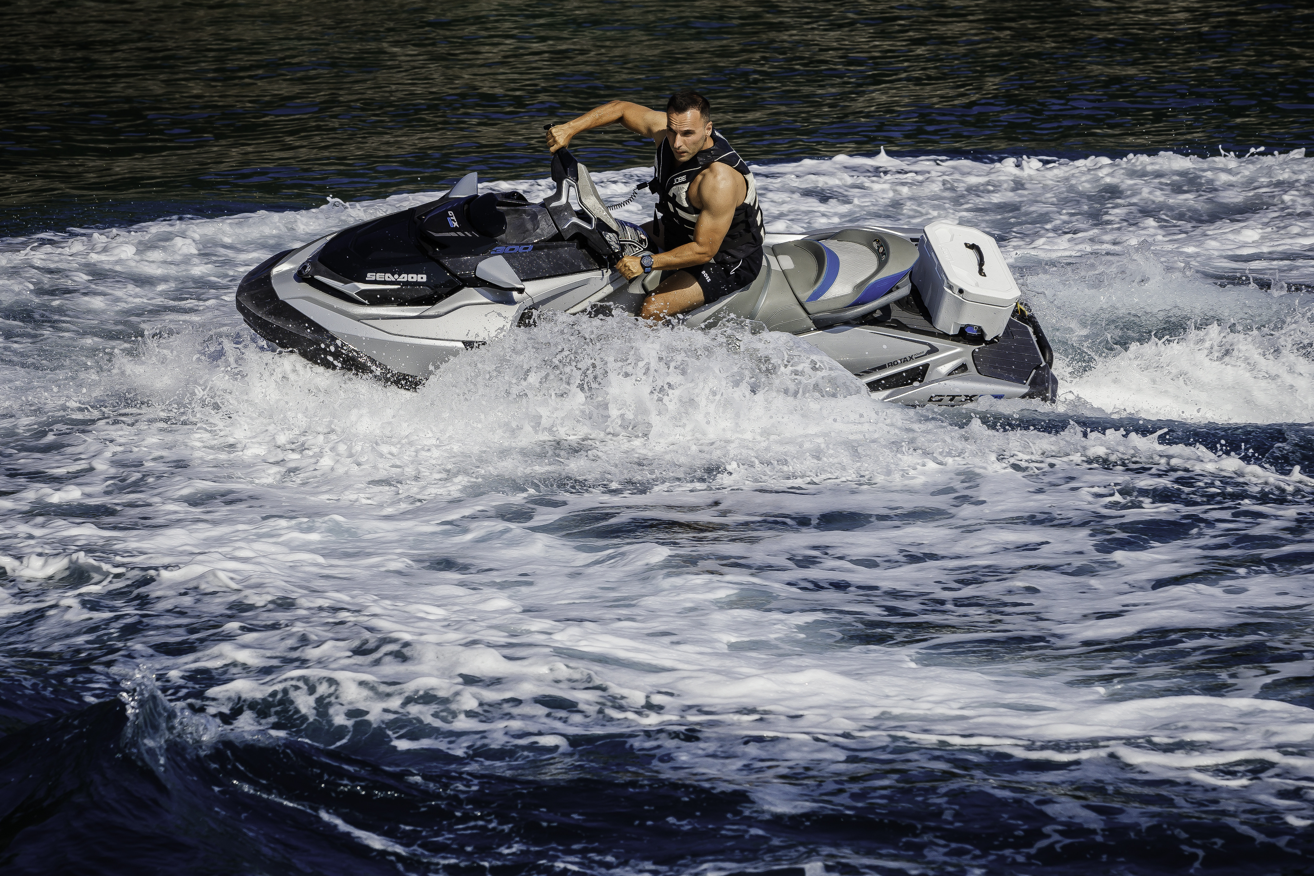 Man riding a jet ski, creating white wake on dark blue water near a coastline.