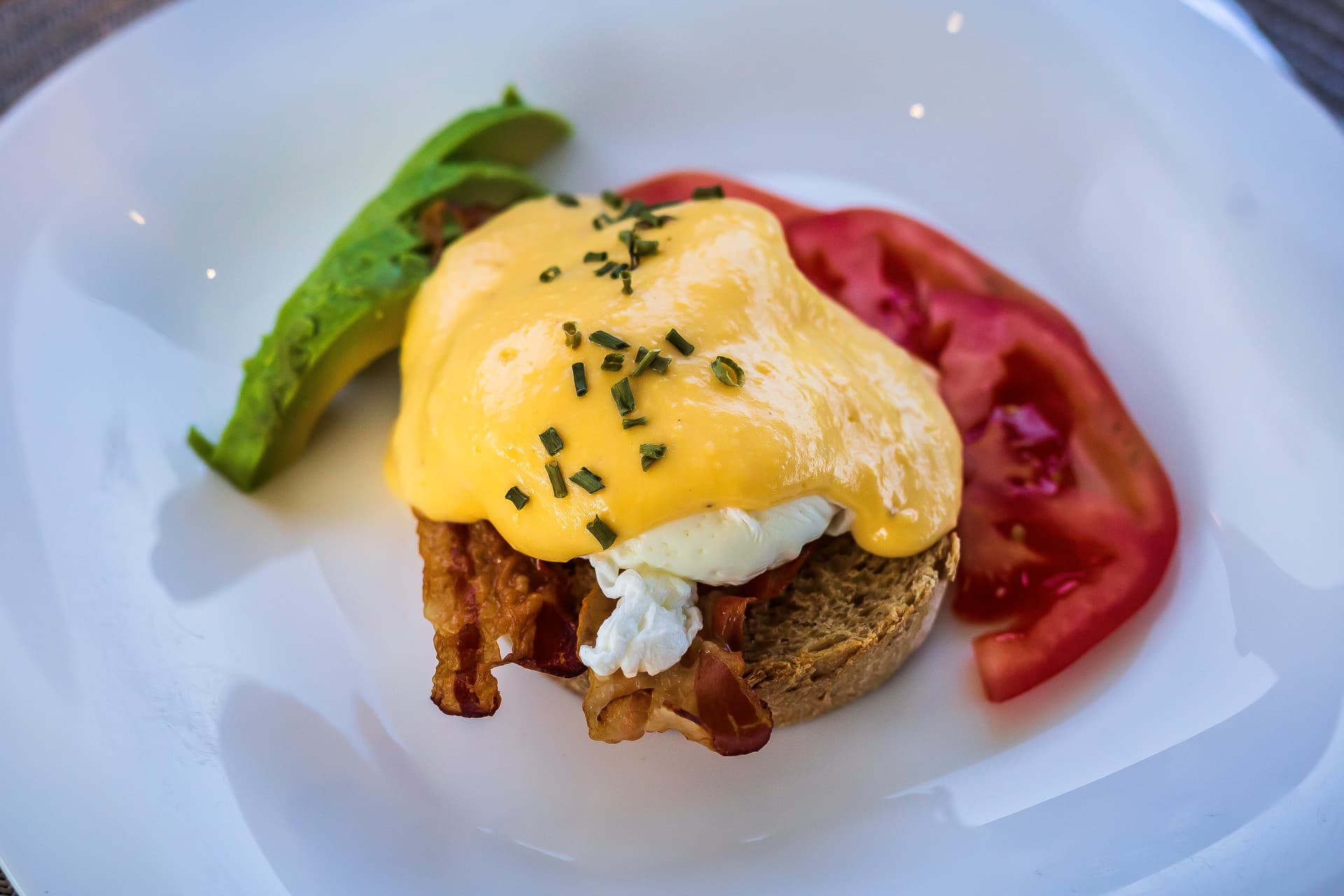 Eggs Benedict with bacon, avocado, and tomato slices on a white plate.