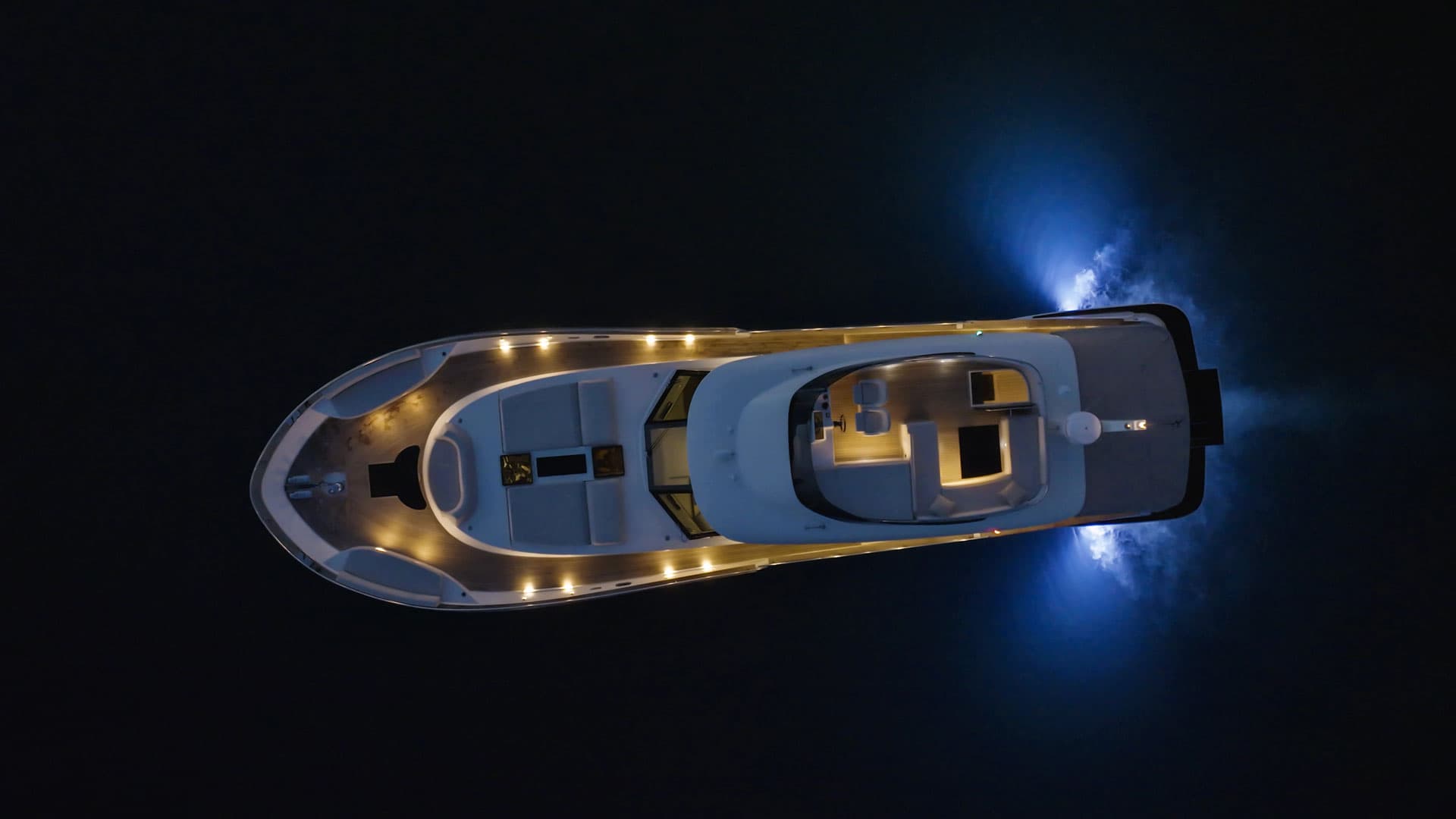 Motor yacht illuminated at night with blue underwater lights creating wake on dark water
