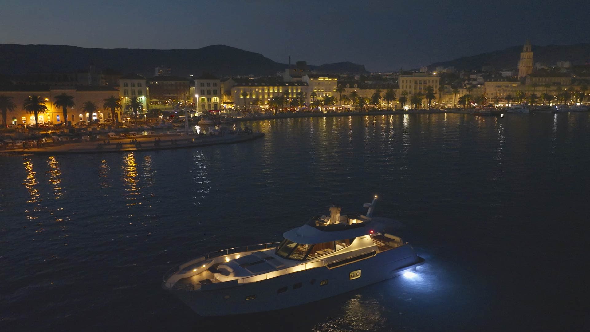 Motor yacht cruising in Split port at night with illuminated waterfront and mountains.