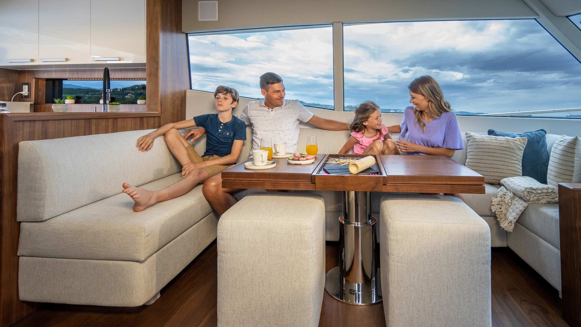 Family dining inside a modern yacht salon with views of the cloudy sky and distant coast.