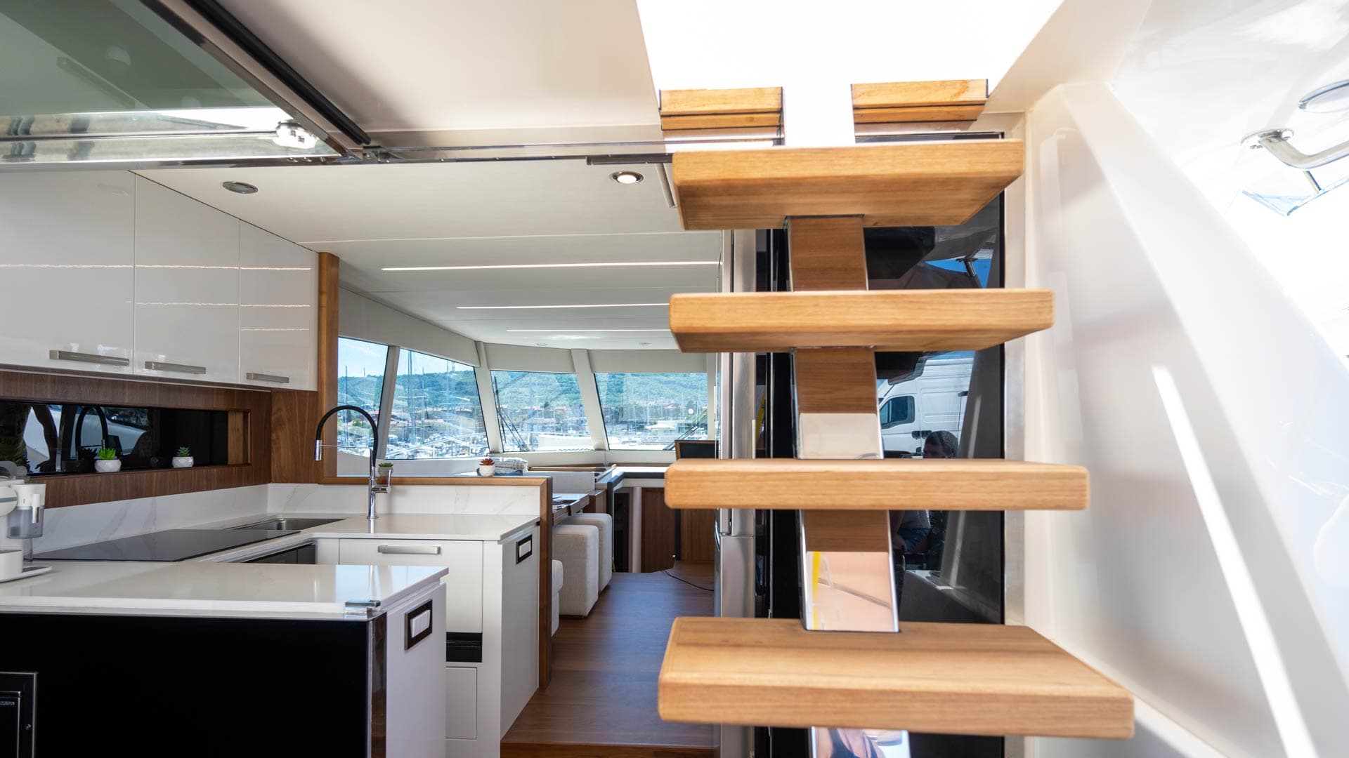 Interior view of a yacht salon with modern white galley, wood accents, and floating stairs overlooking a marina.