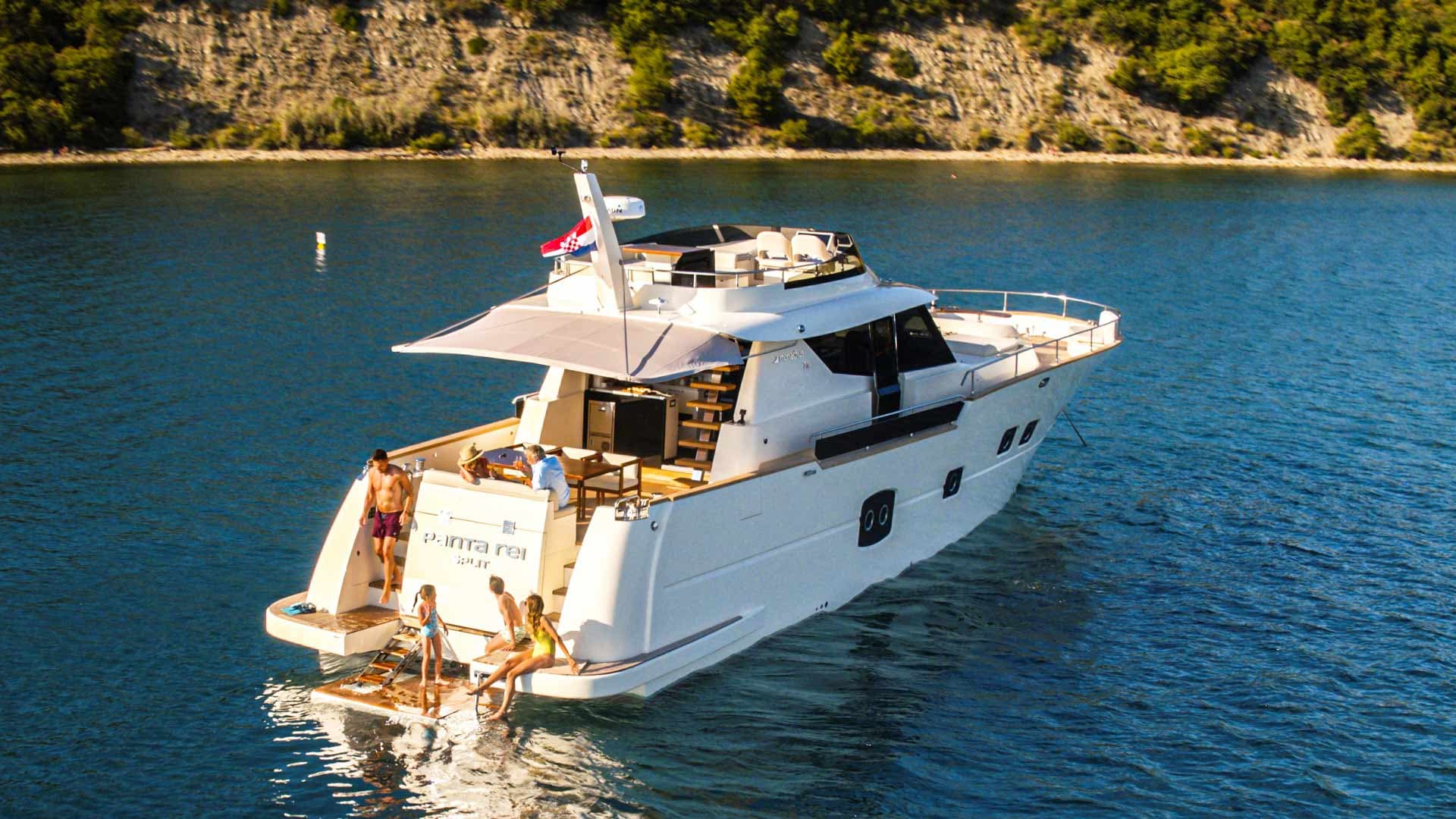 Motor yacht "PANTA REI" with people relaxing near the stern on blue coastal waters near a green cliff.