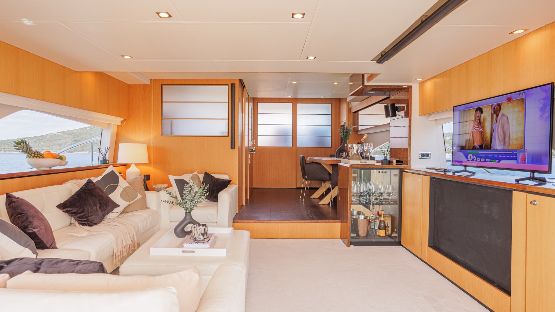 Yacht salon interior with white leather seating, wood paneling, and view of coastline.
