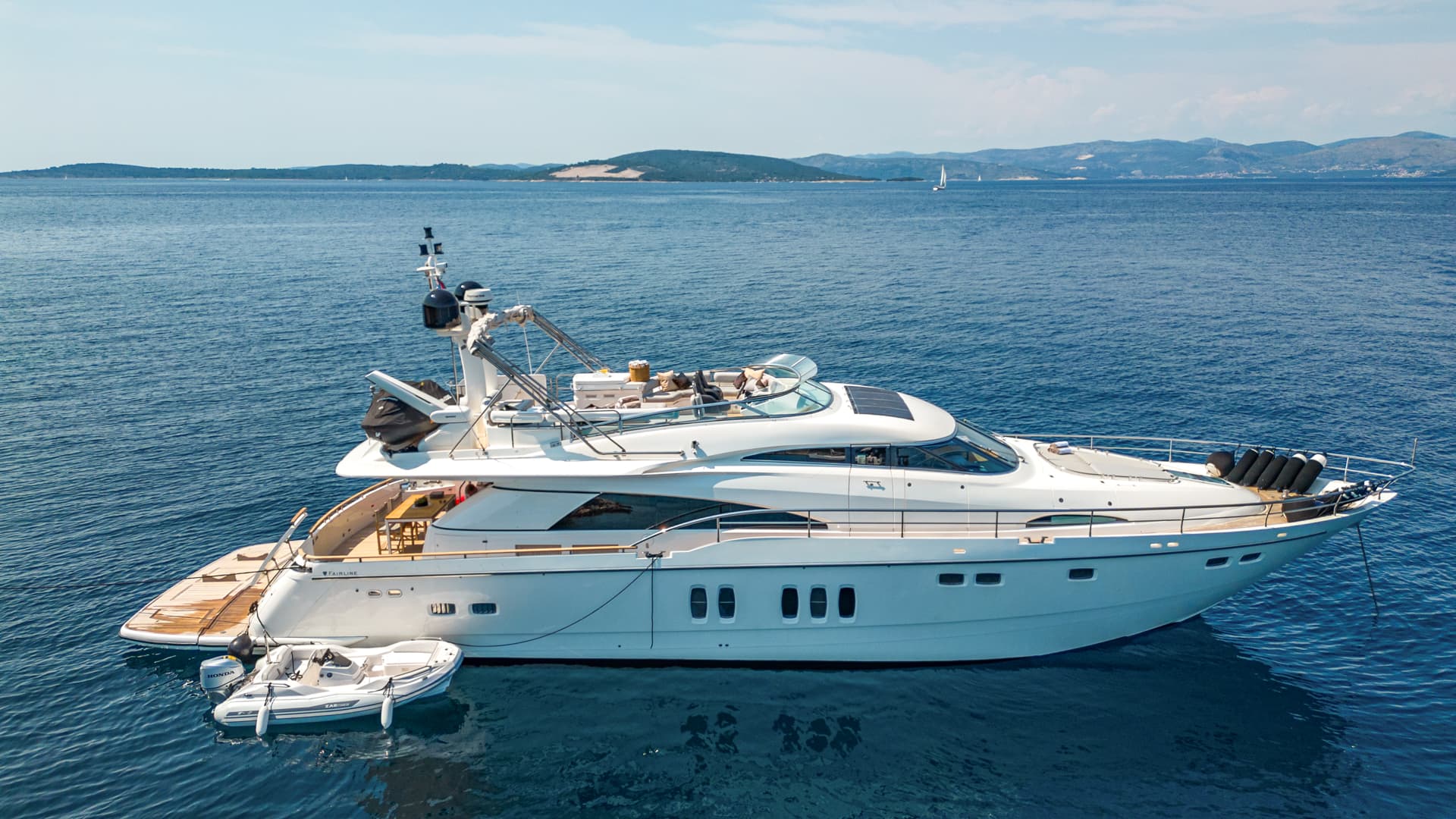 Large white motor yacht anchored on blue sea near hilly coastline with distant sailboats.