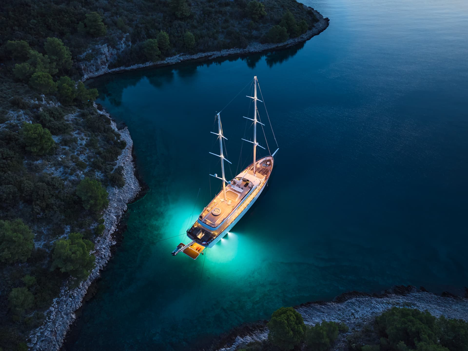 Large sailboat anchored in dark cove with illuminated turquoise water and forested coastline.