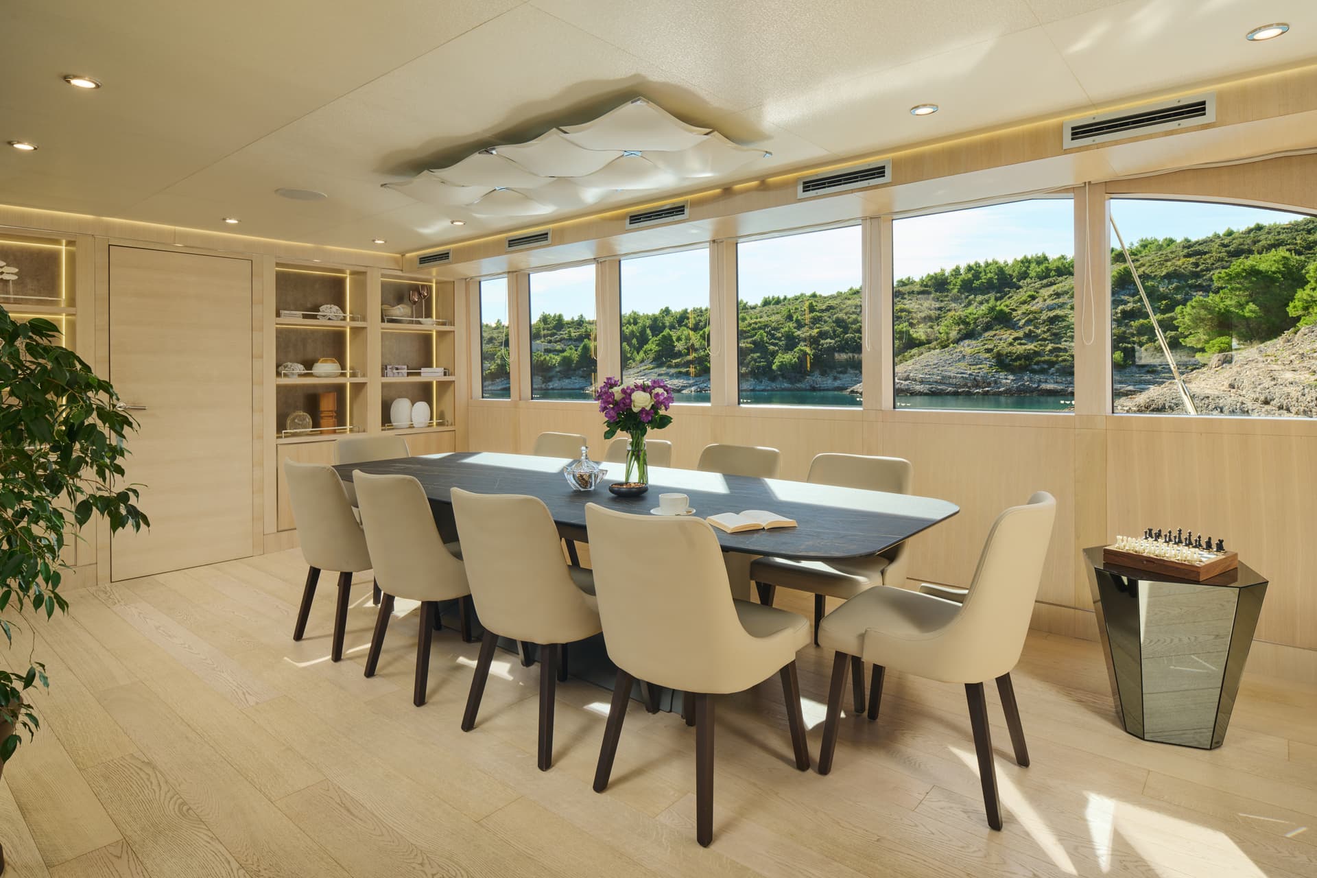Yacht dining room with large windows overlooking a lush, rocky coastline and blue water.