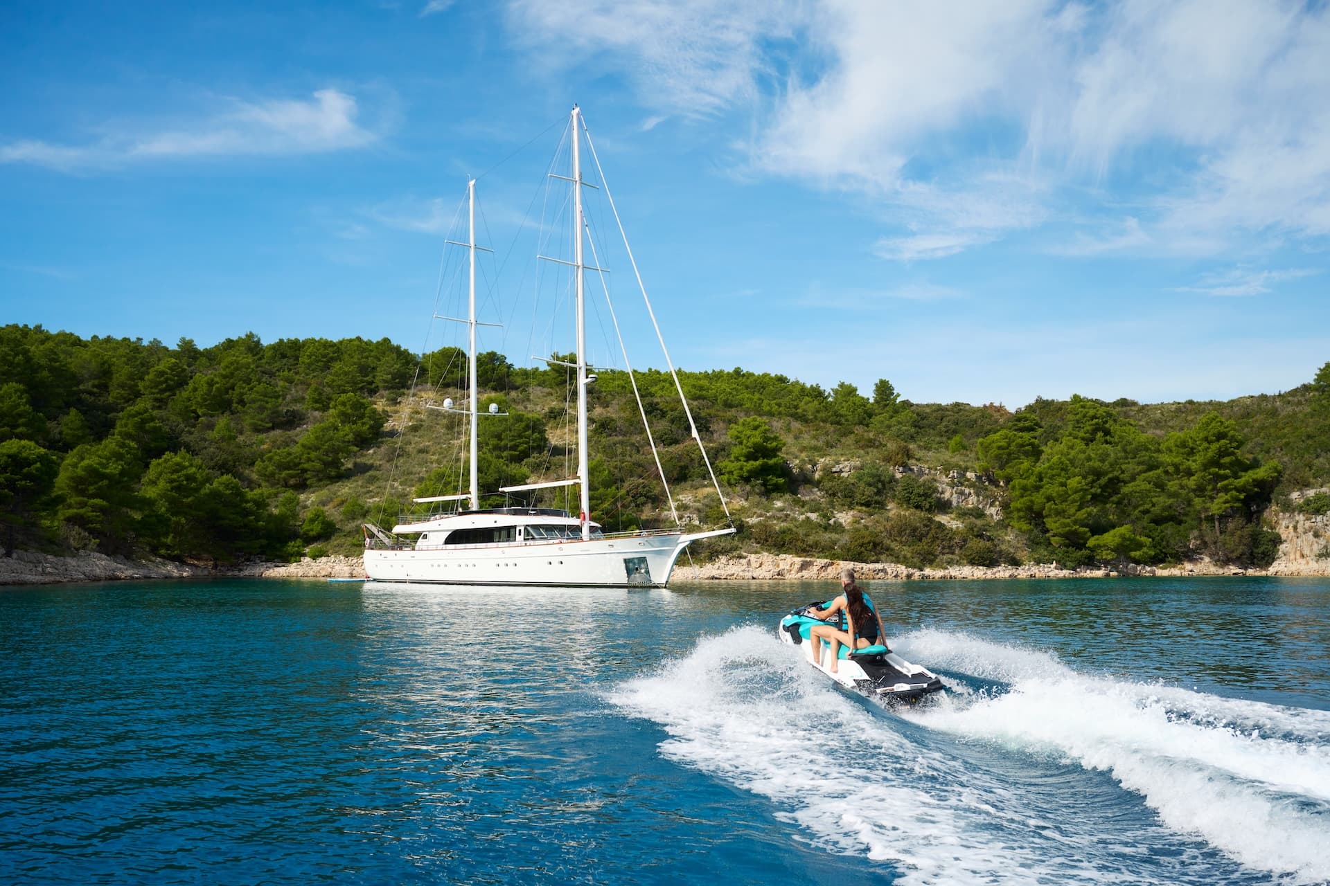 Woman riding a jet ski past a large white sailboat anchored near a wooded coastline.