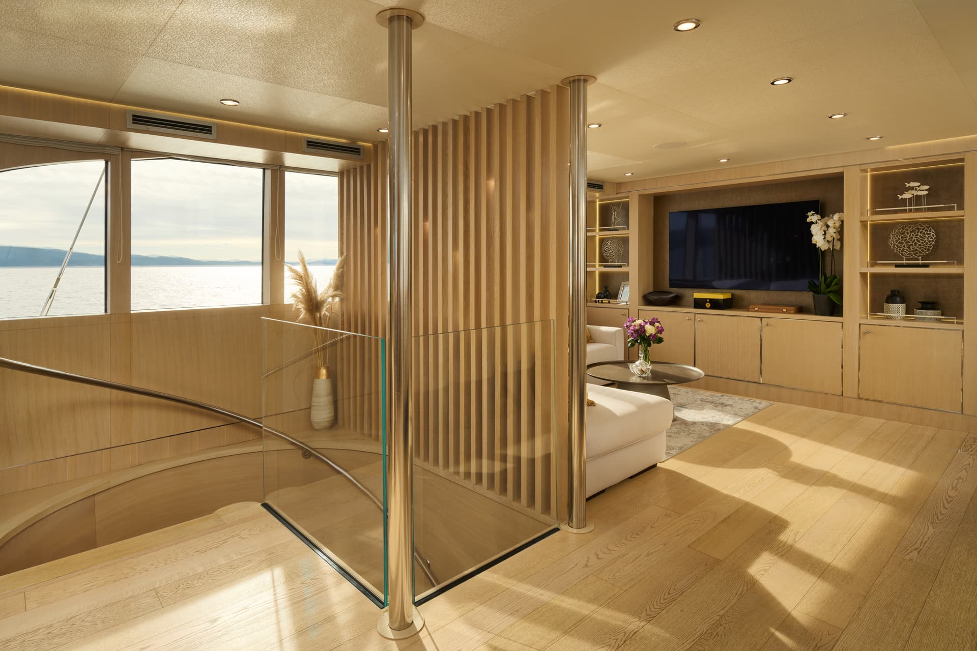 Luxury yacht interior with light wood, glass railing, and ocean view of mountains.