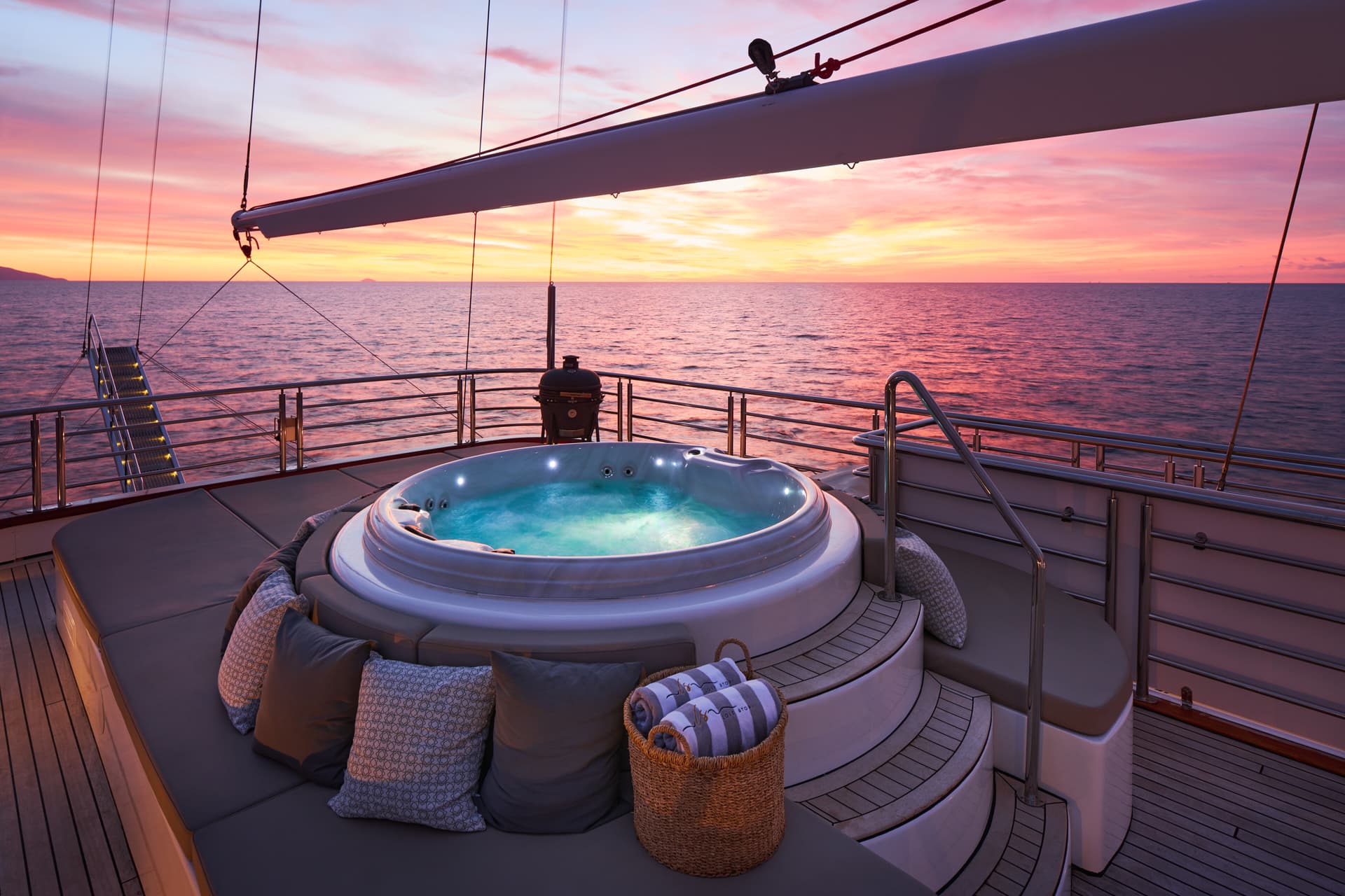 Hot tub on yacht deck with cushions and towels against a vibrant pink and orange sunset over the sea.