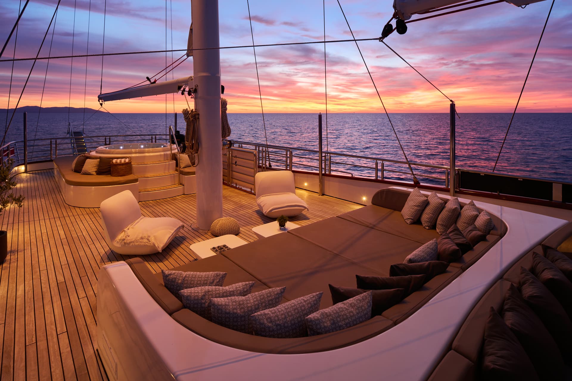 Yacht deck with lounge seating and hot tub overlooking ocean at vibrant sunset with distant mountains.