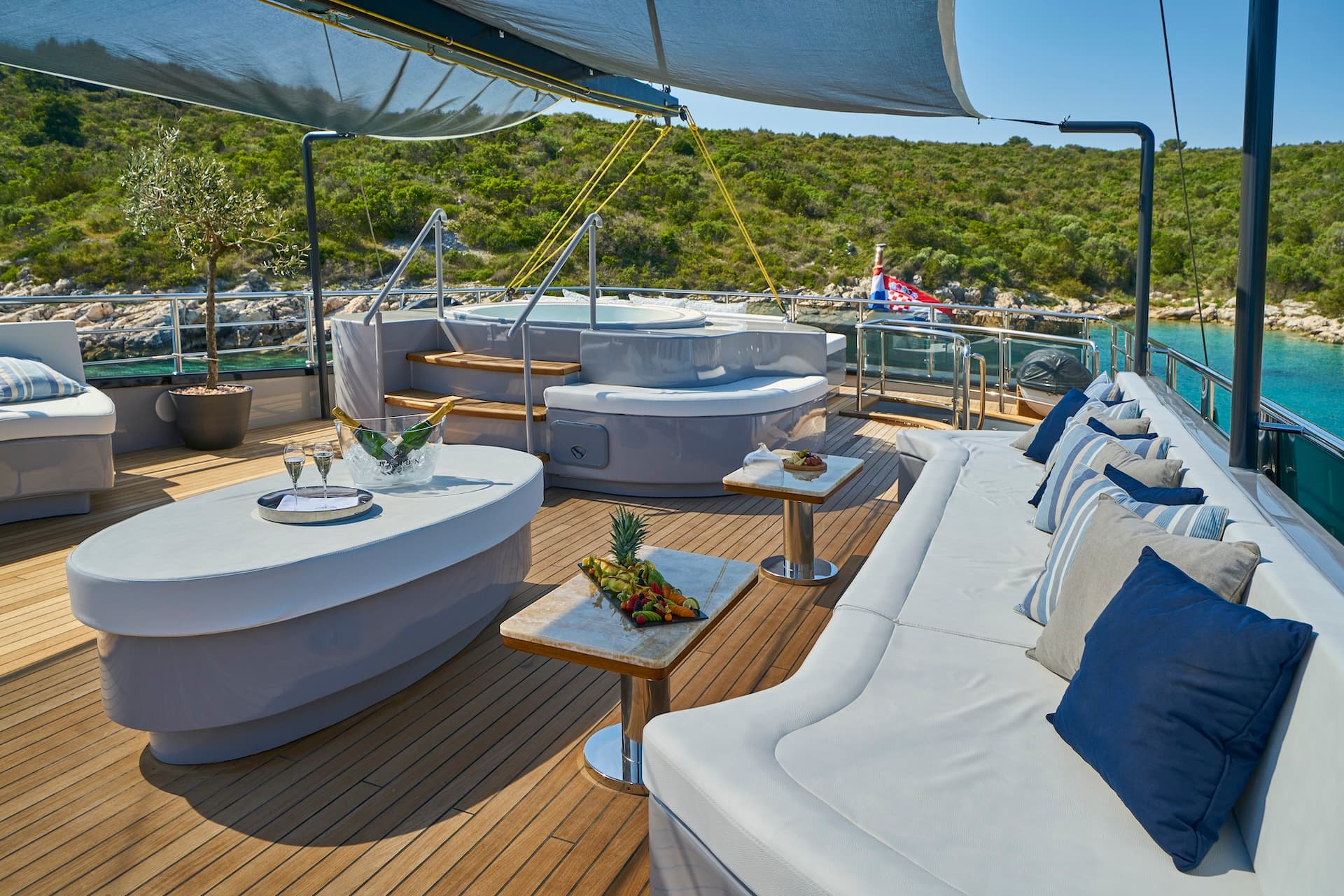 Luxury yacht deck with hot tub, champagne, and fruit platter anchored near green coastline.