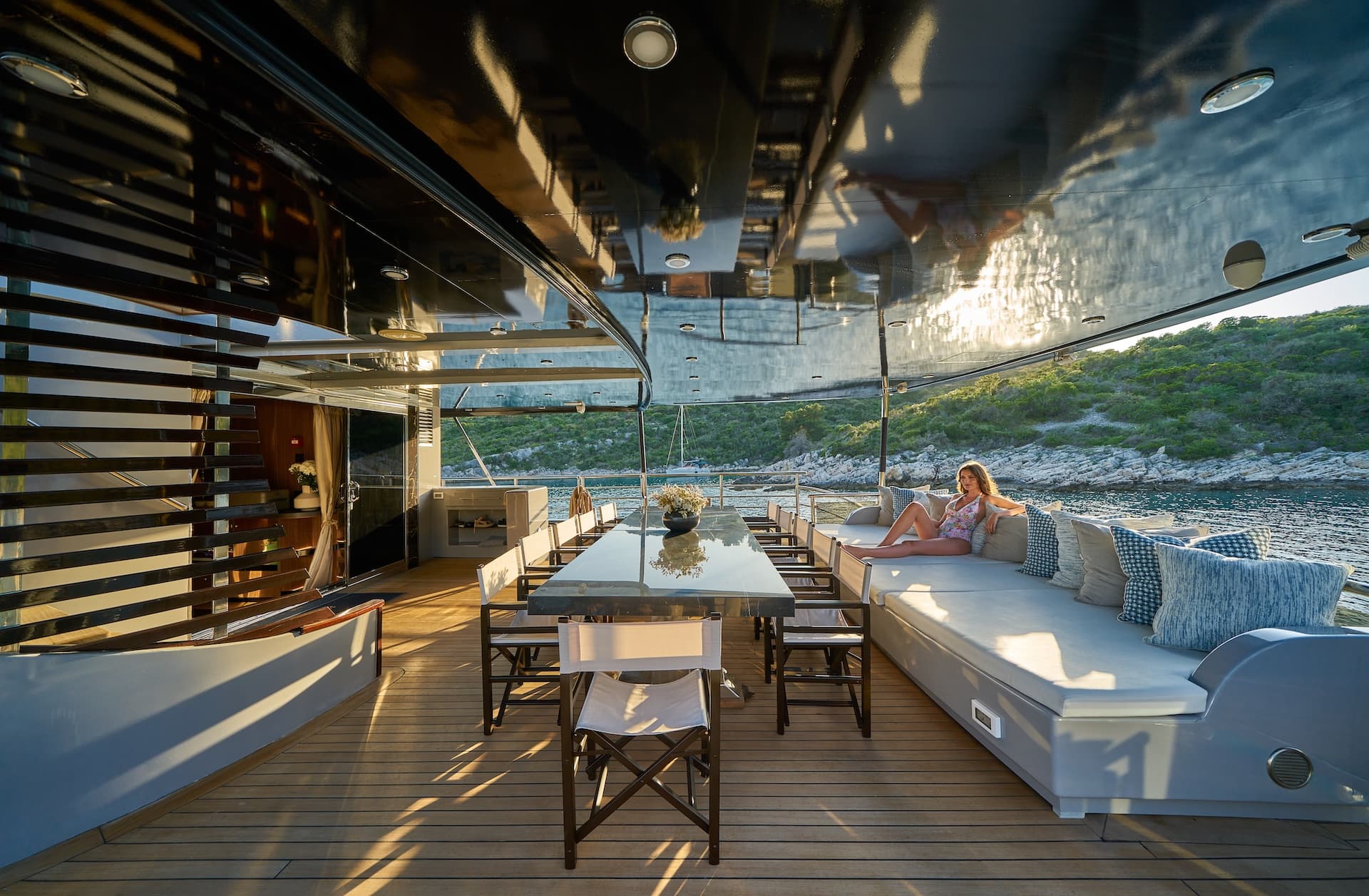 Luxury yacht deck with dining table, sun lounge, and woman relaxing near a green, rocky coastline.
