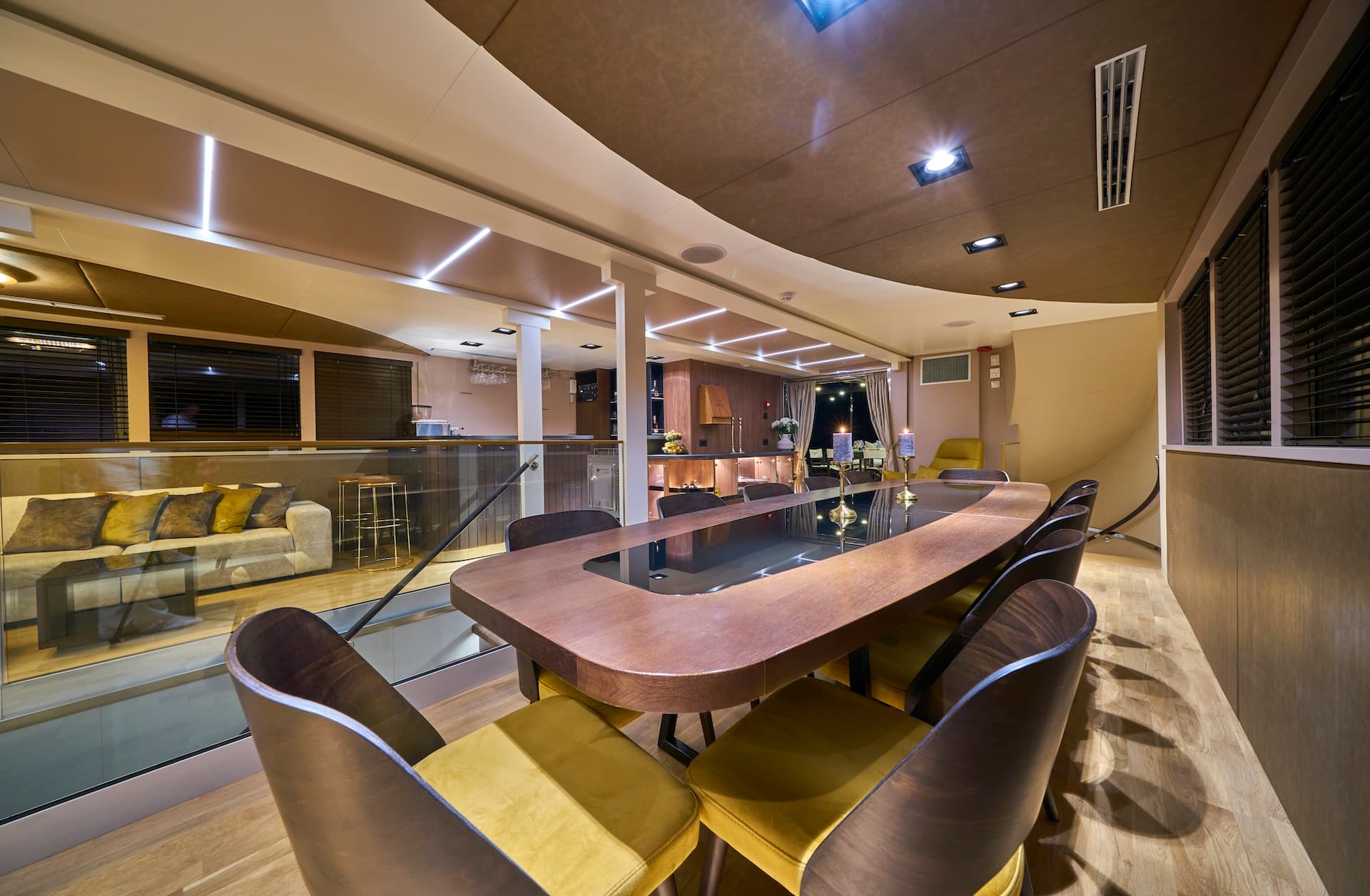 Luxury yacht dining area with wood table, yellow chairs, and sunken lounge visible below.