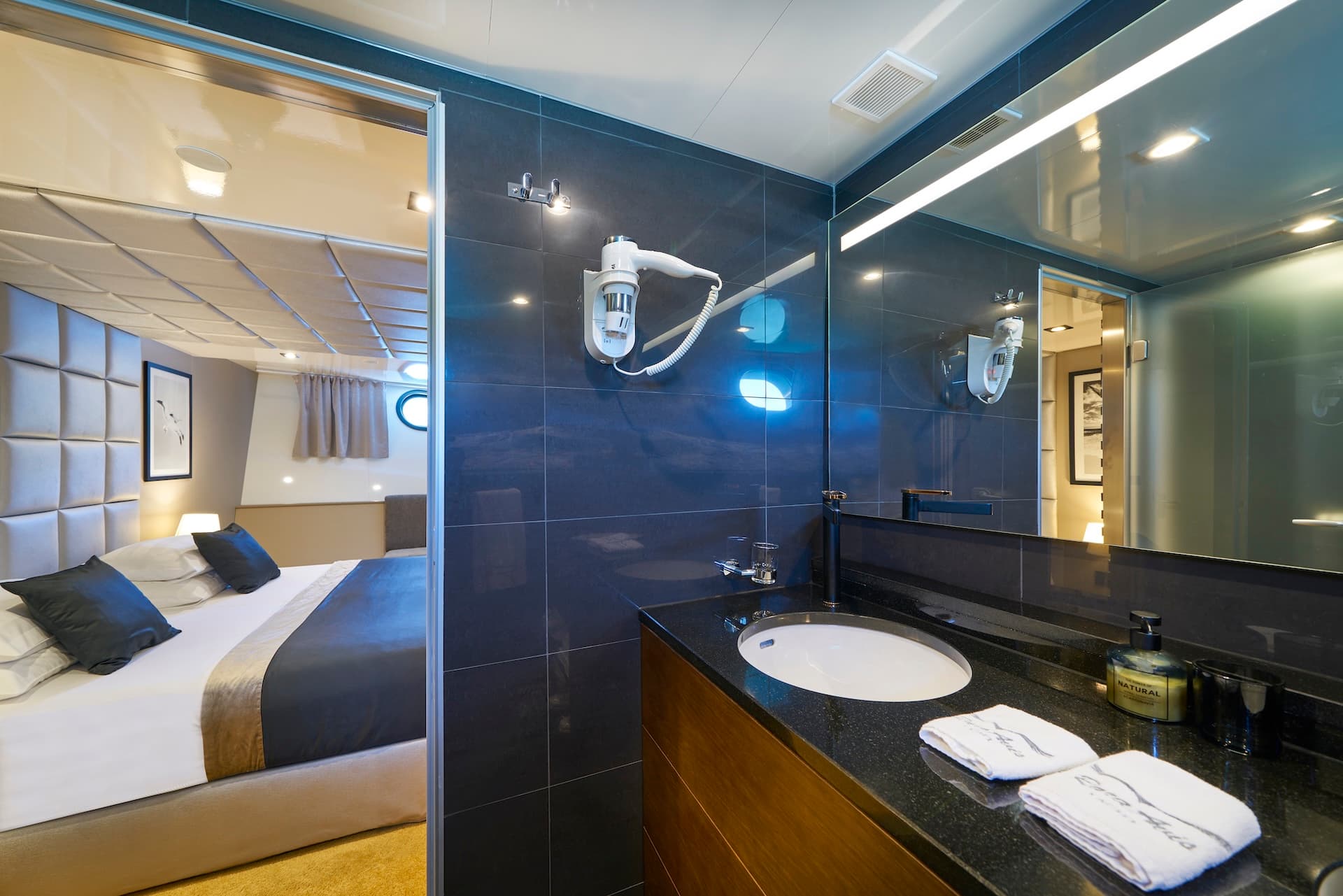 Yacht cabin bathroom with dark tile and view into modern bedroom with porthole window.