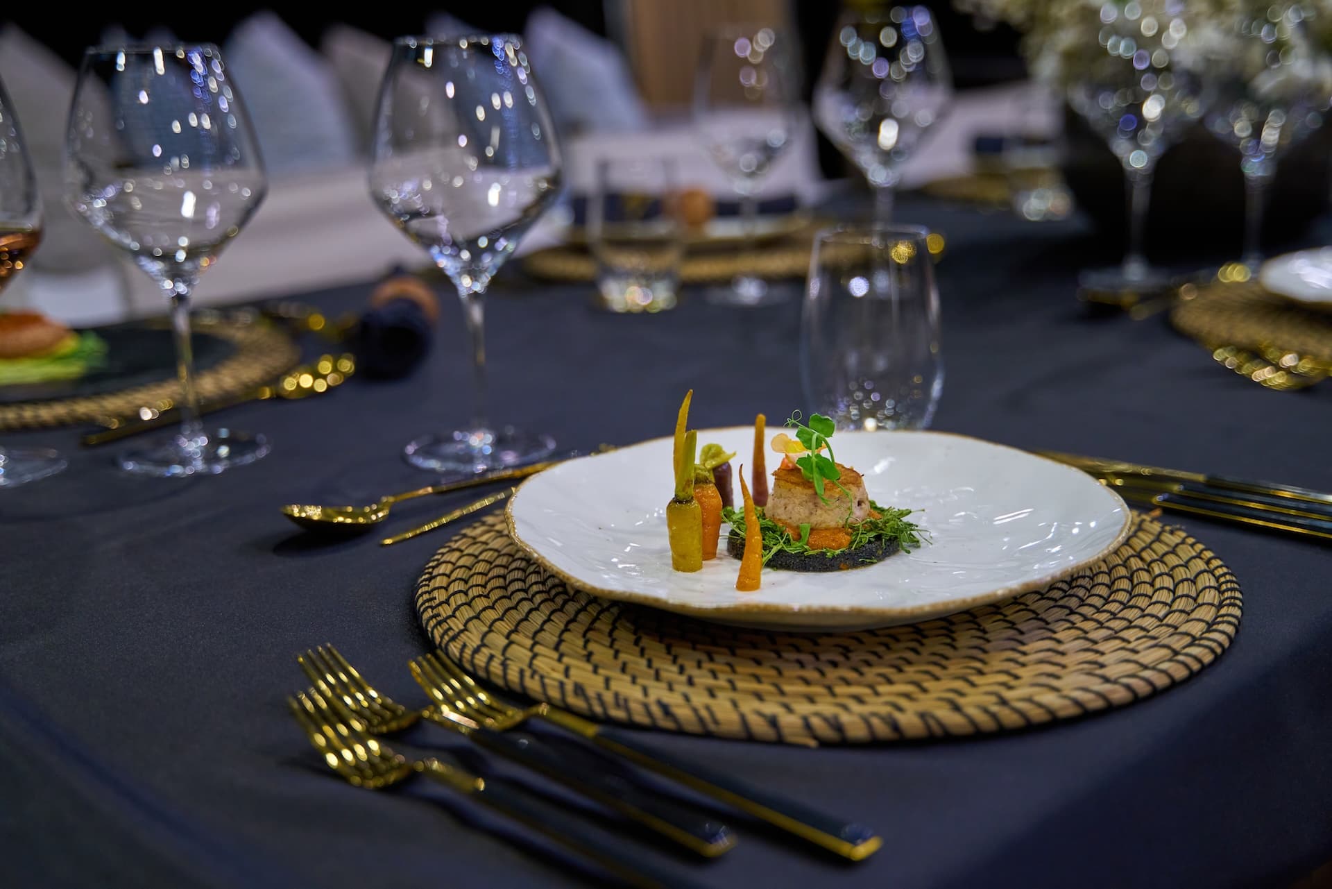 Gourmet appetizer on white plate with gold cutlery and wine glasses on dark tablecloth.