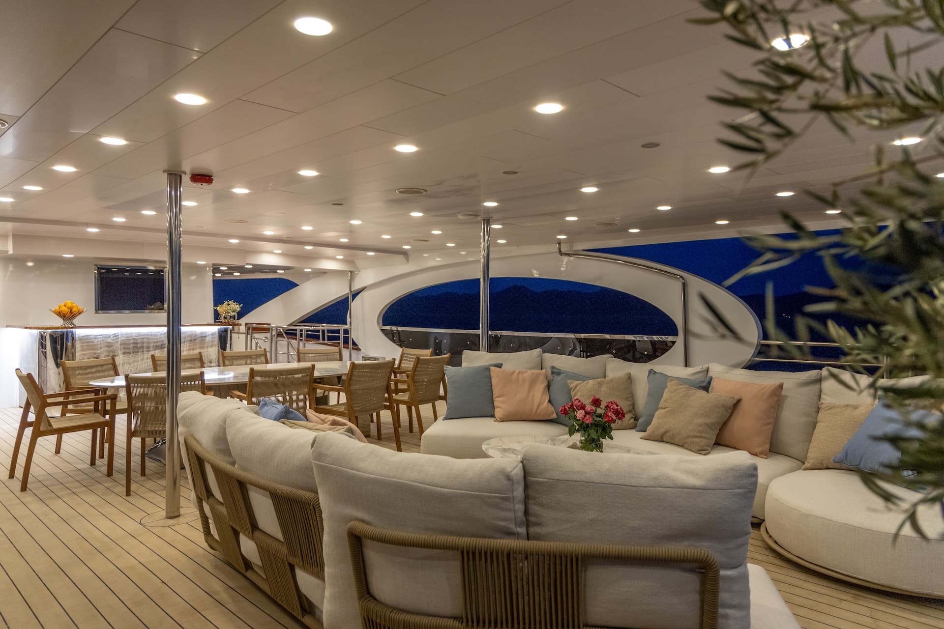Luxury yacht deck lounge with white seating, dining area, and dark mountains visible outside.