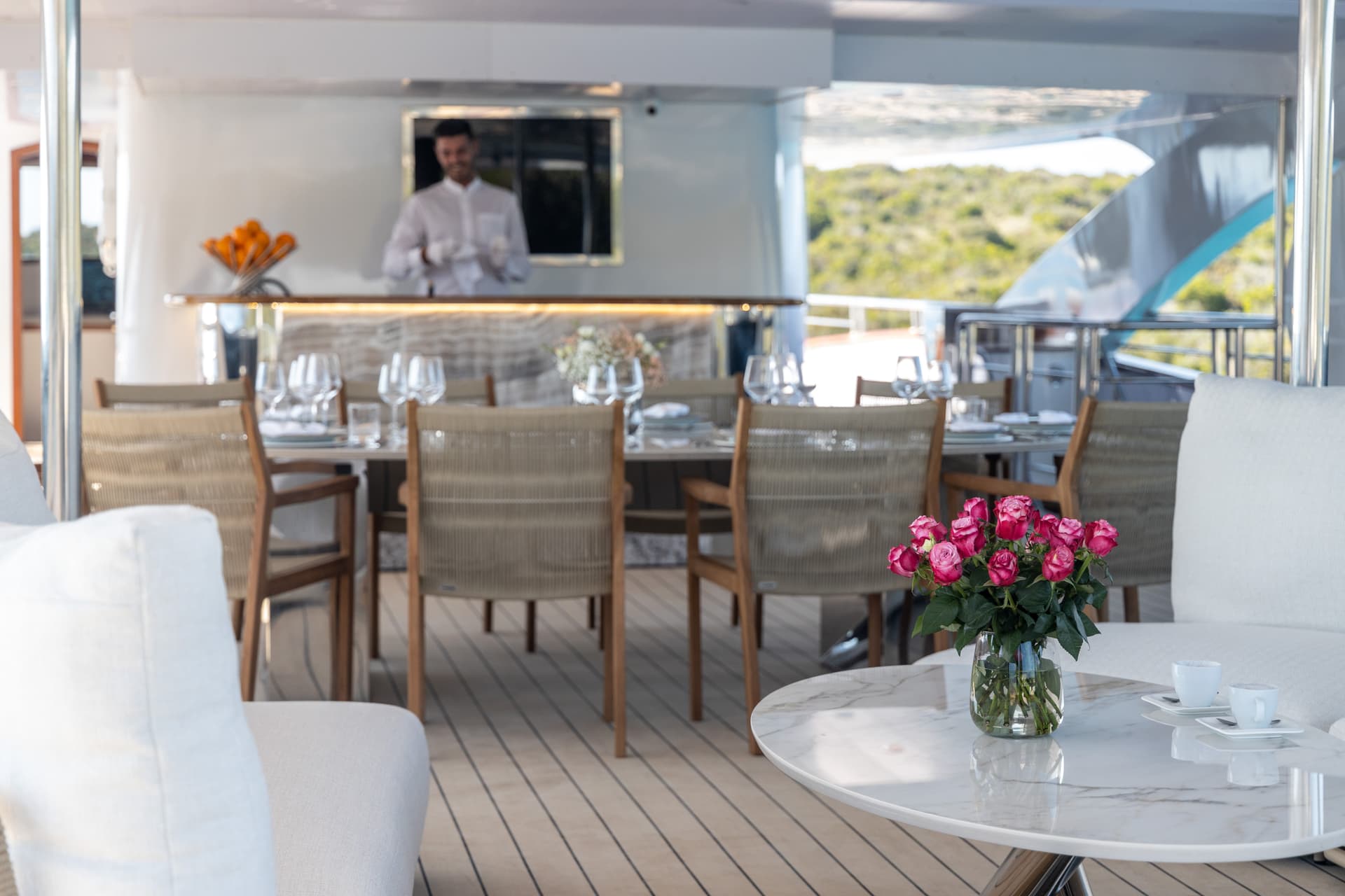 Outdoor dining area on a yacht deck with a bartender serving and island scenery.