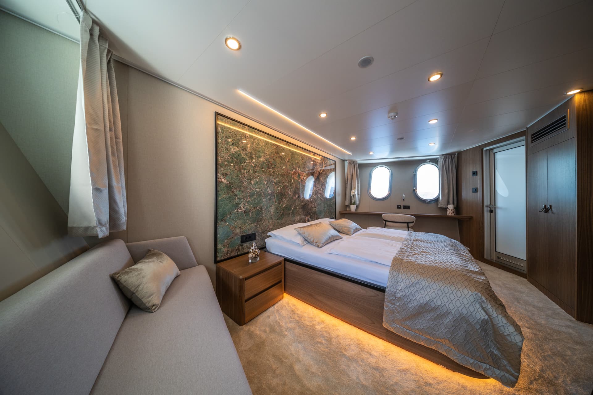 Modern yacht cabin bedroom with porthole windows and illuminated green marble headboard.
