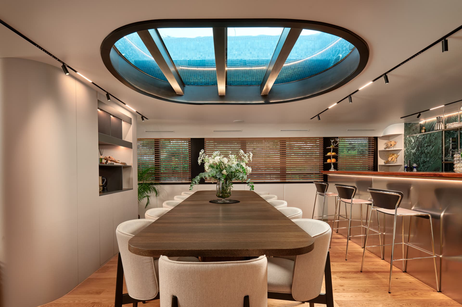Modern yacht dining area with long wooden table, upholstered chairs, and oval skylight showing water.