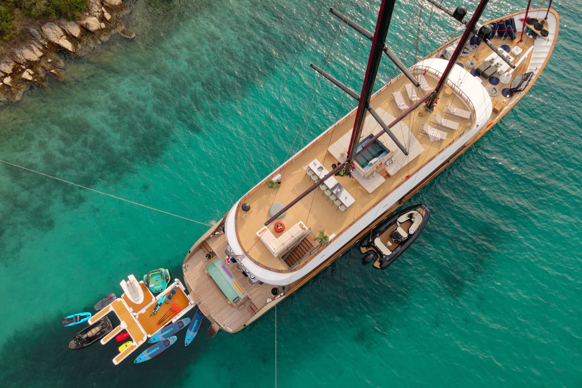 Large yacht anchored near rocky shore with turquoise water and water toys.