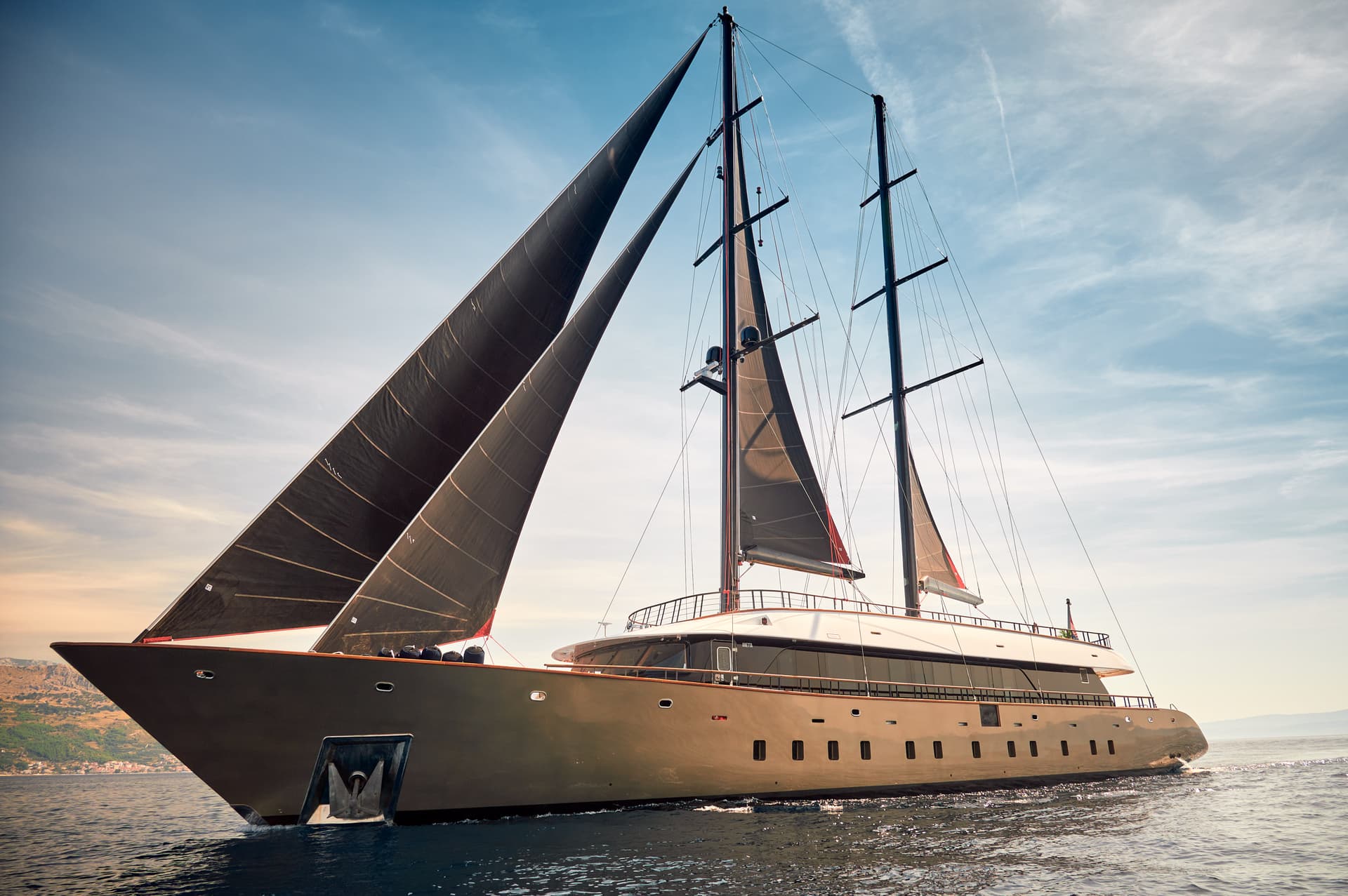 Large luxury sailing yacht Anetta with black sails on coastal waters near a hilly shore.