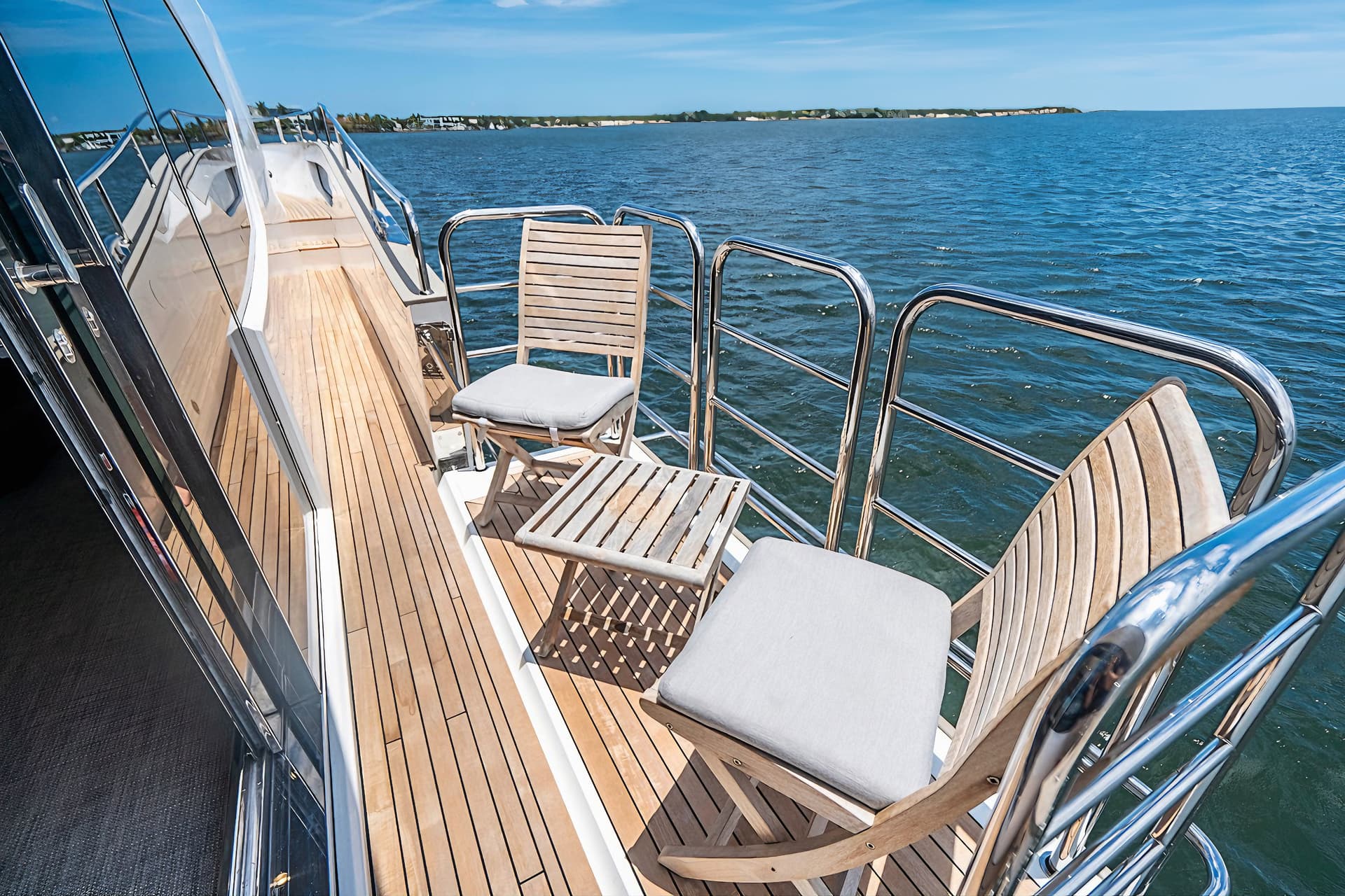 Aft deck of a yacht with teak flooring, wooden chairs, and chrome railings on blue water.