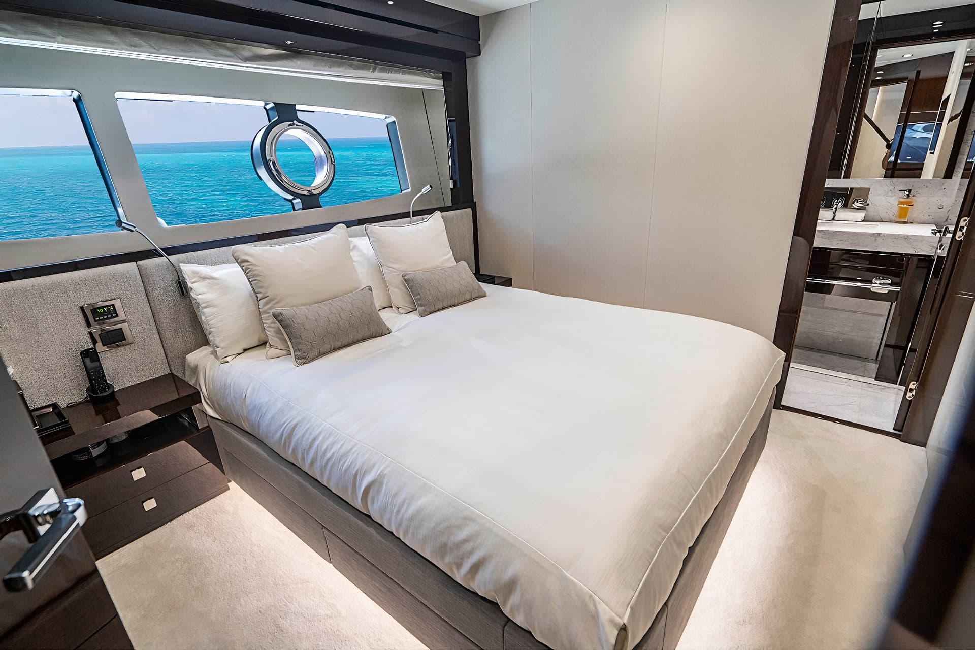Yacht cabin bedroom with white bedding overlooking turquoise ocean through rectangular and porthole windows.