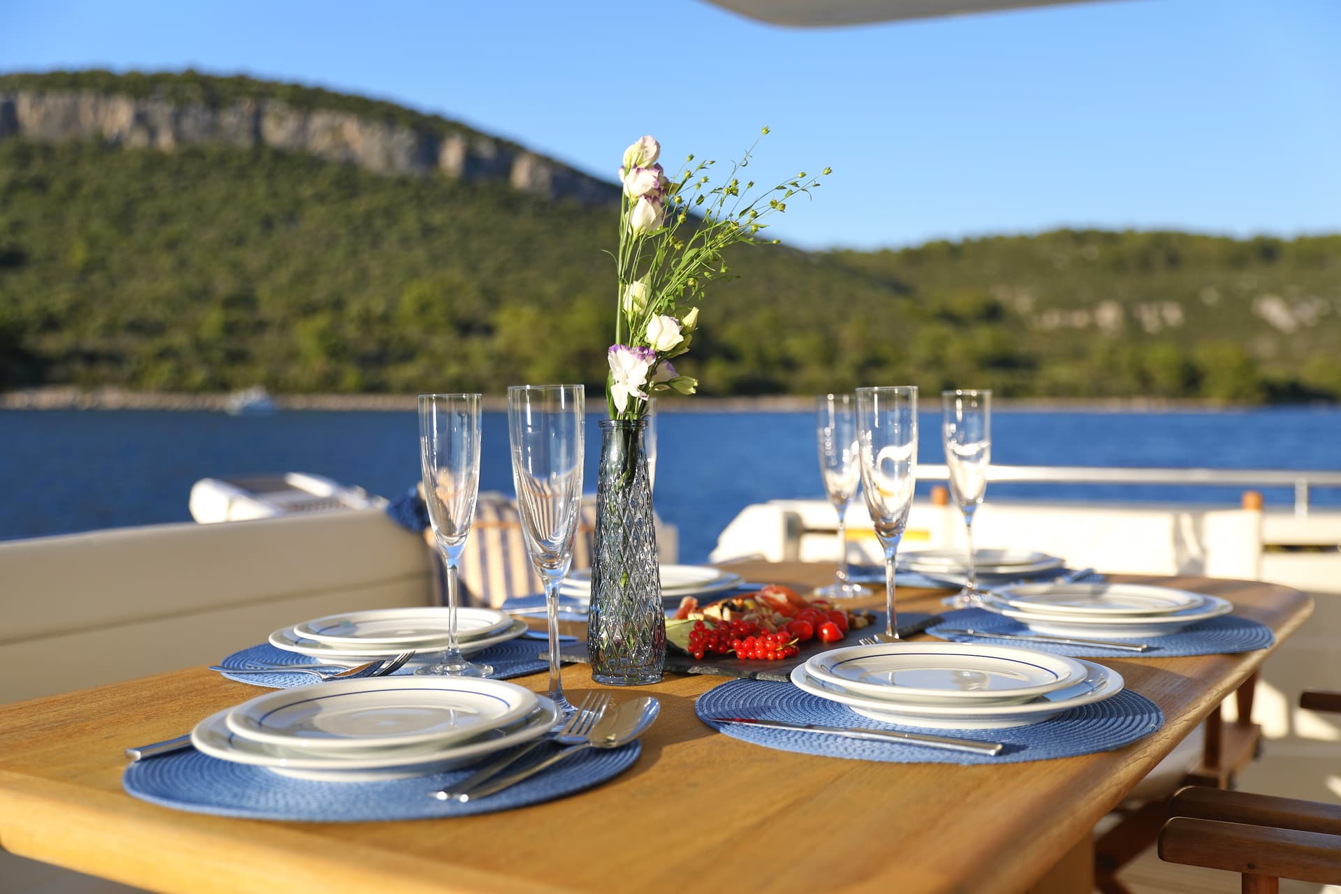 Table set for dining on a yacht deck with a floral centerpiece and coastal mountains in the background.