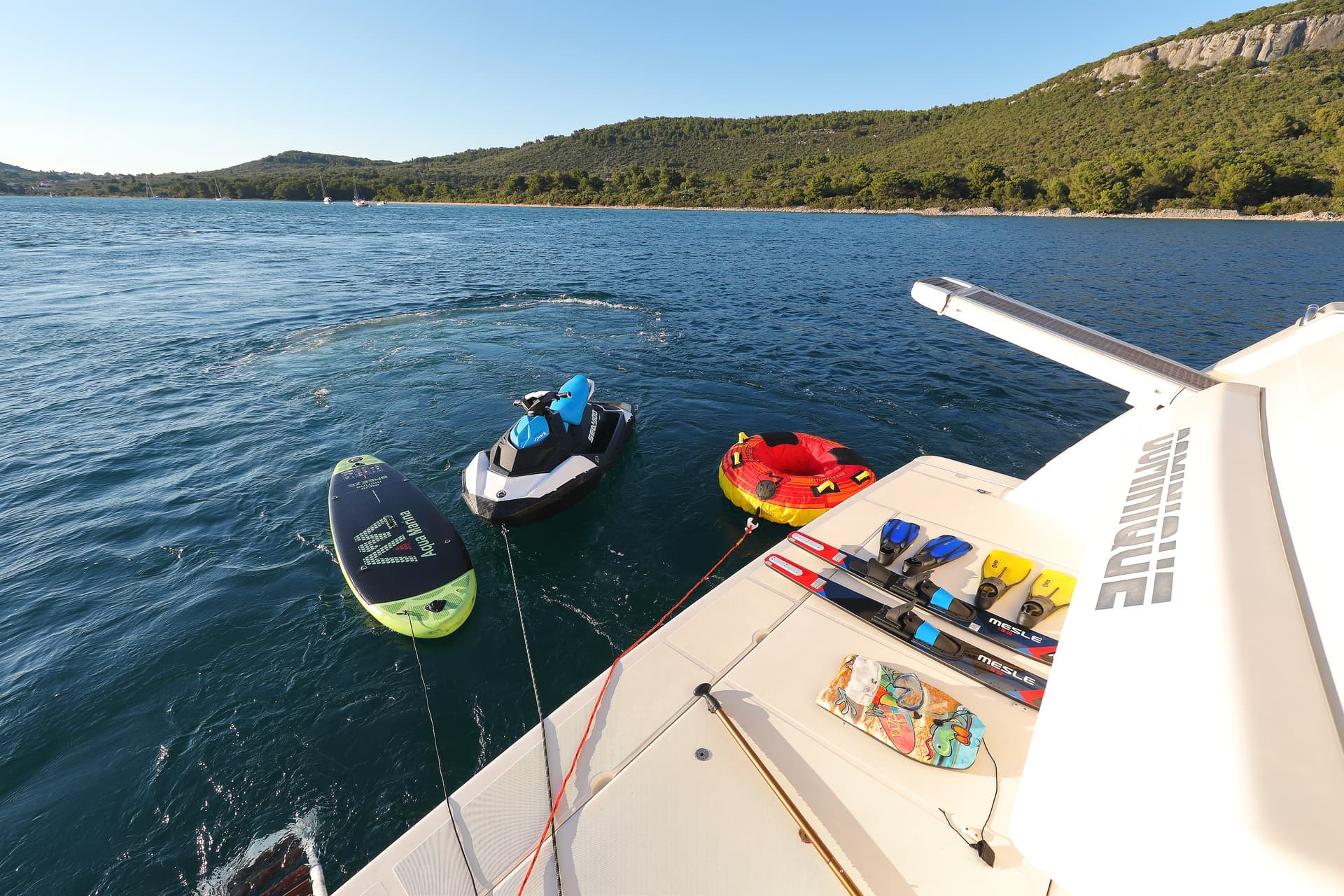 Water toys, jet ski, and skis on a yacht deck near a wooded coastline.