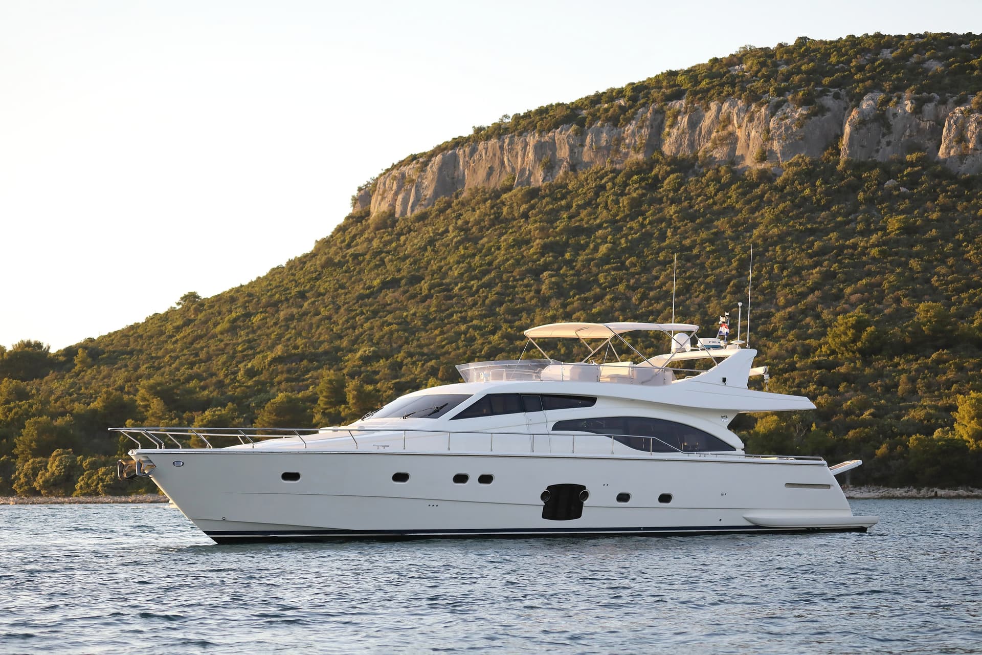 White motor yacht anchored near a steep, forested coastline under bright sky.