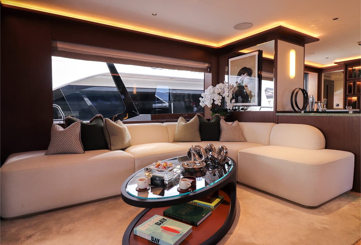 Yacht saloon with cream sectional sofa, wood paneling, and view of another boat.
