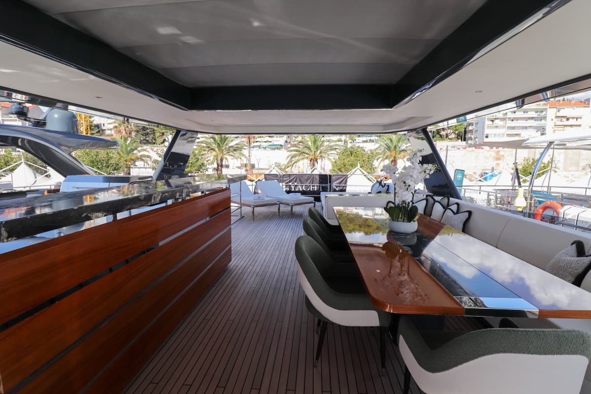 Flybridge dining area on a yacht with teak deck, bar, and palm trees ashore.