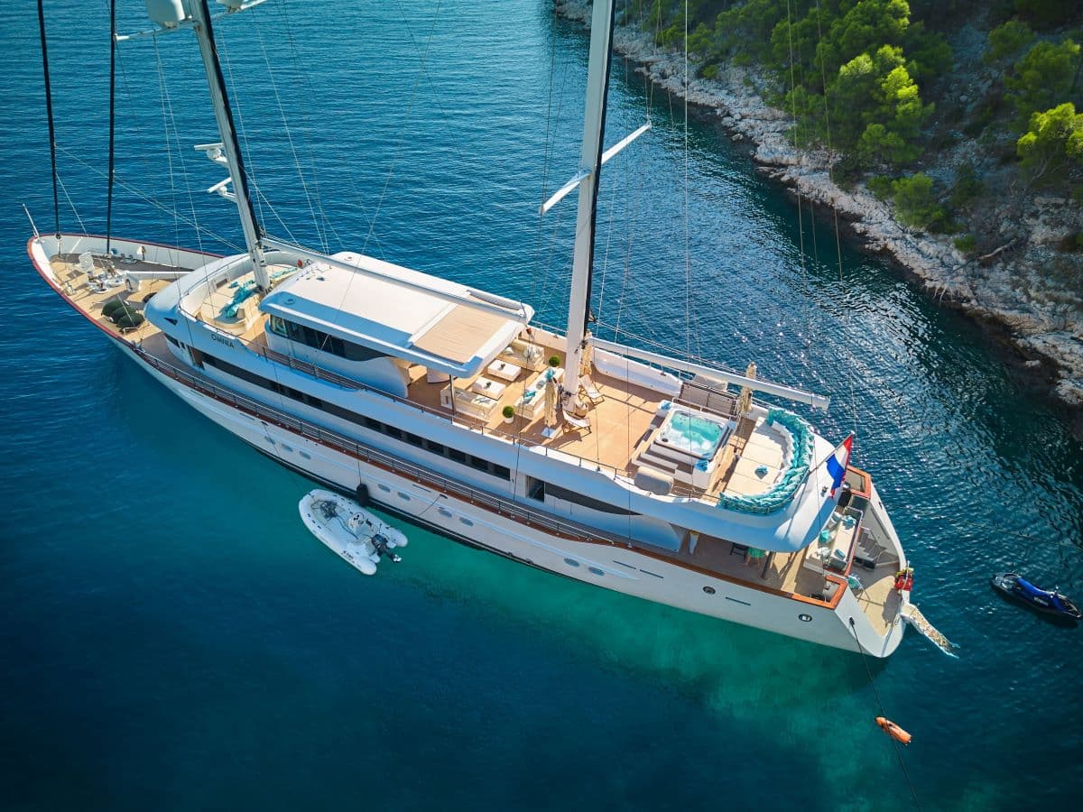 Large white sailing yacht anchored near a rocky, tree-lined coast in clear blue water