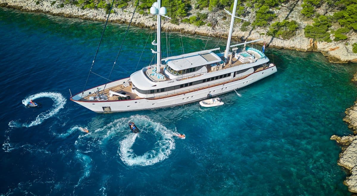 Large white yacht anchored near rocky, tree-lined coast with people riding personal watercraft.