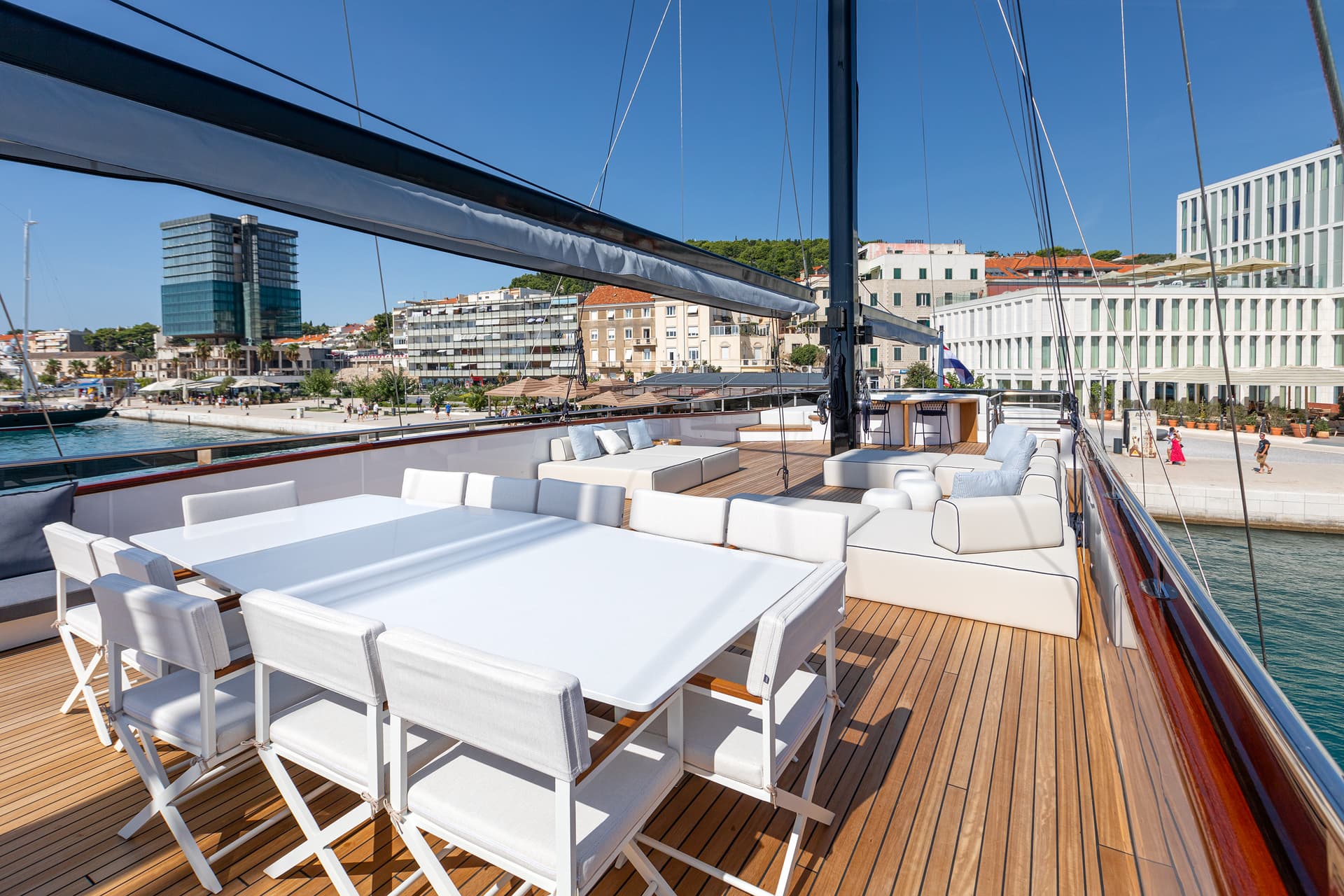 Yacht deck with white dining set and lounge seating overlooking a sunny harbor city.