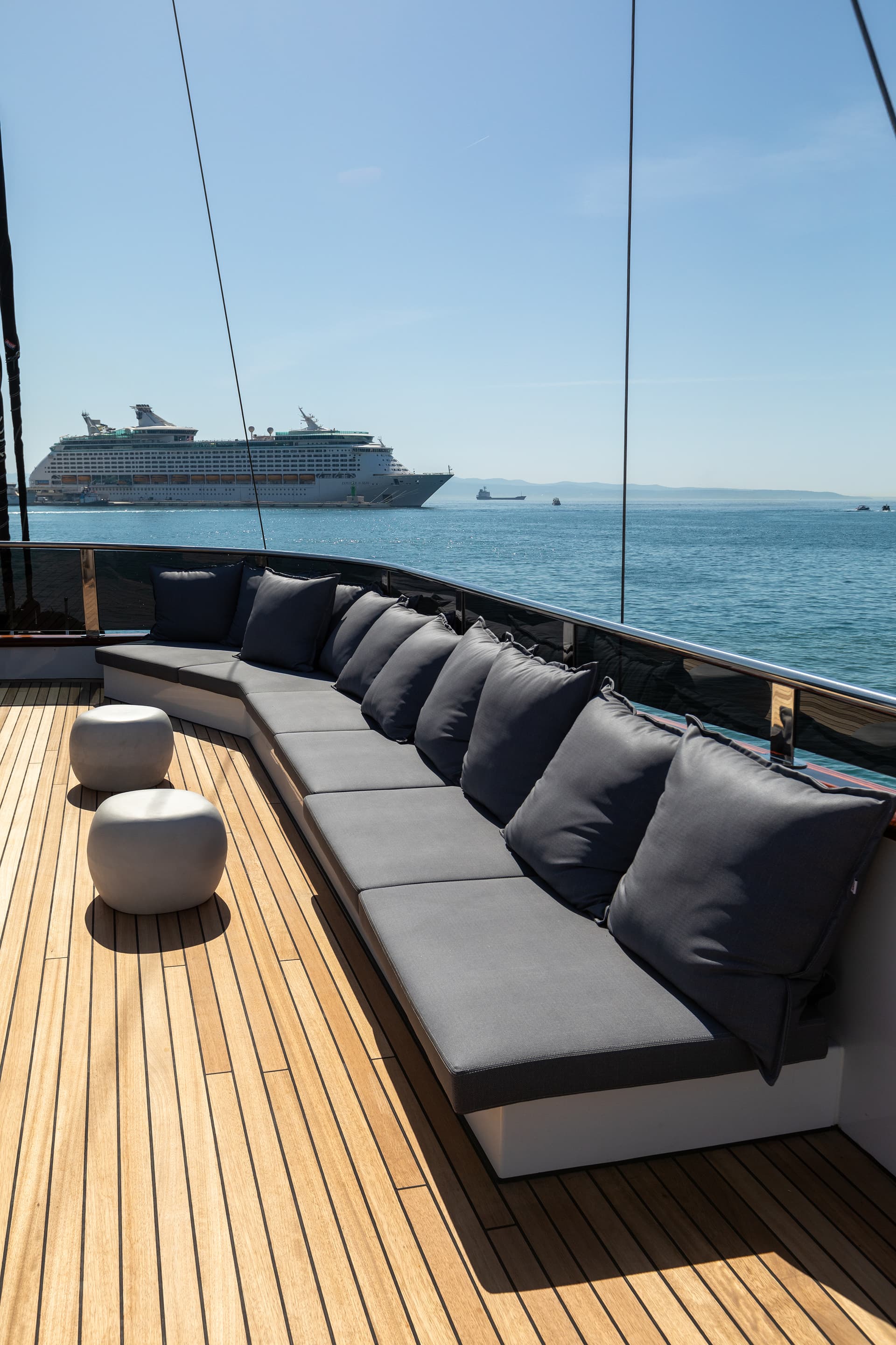 Teak deck seating area with grey cushions overlooking blue sea and a large cruise ship.