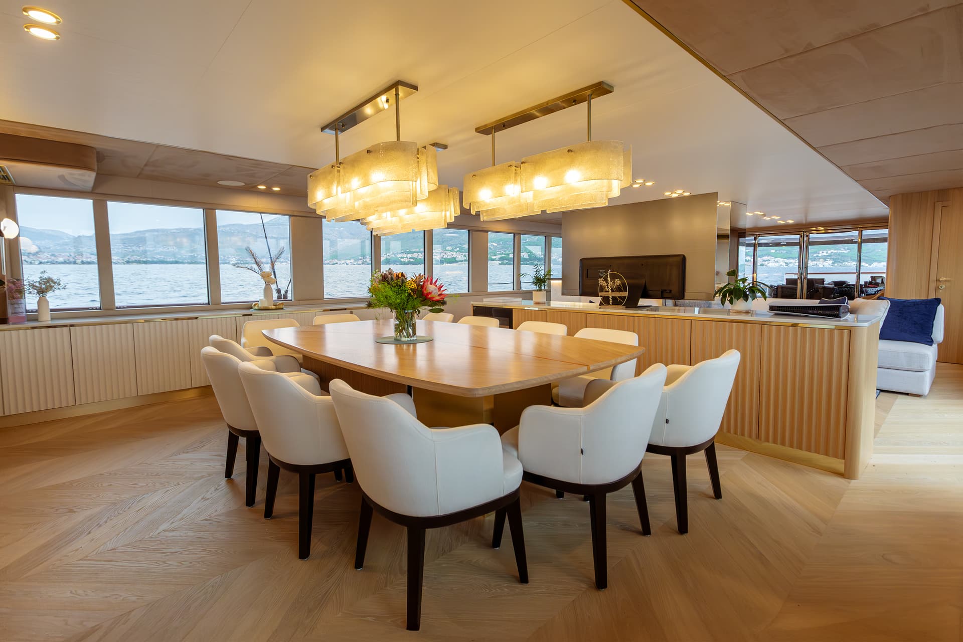 Luxury yacht dining area with white chairs, wood table, and sea view of mountains.