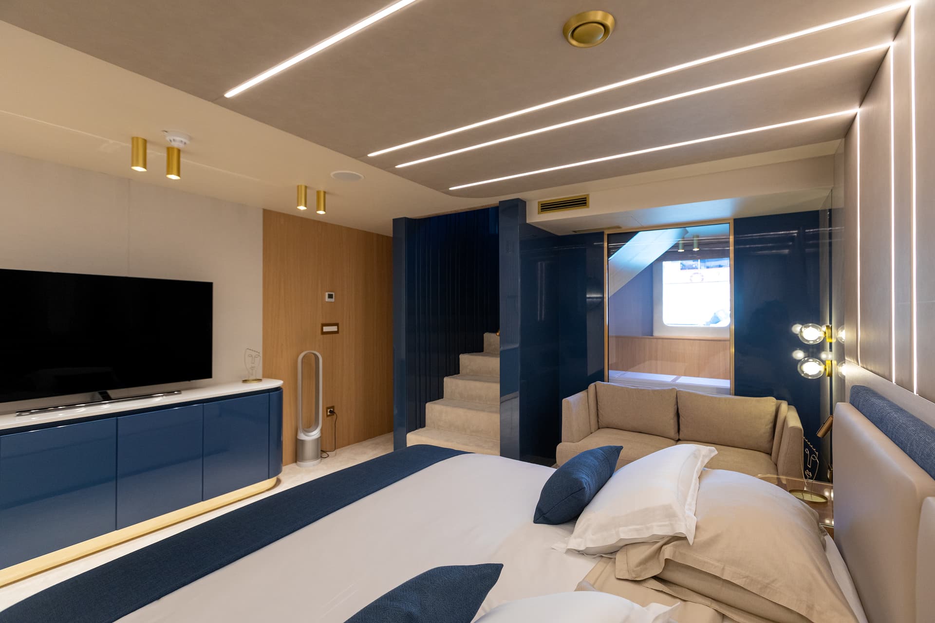 Modern yacht cabin interior with blue accents, plush bedding, TV, and stairs to upper level