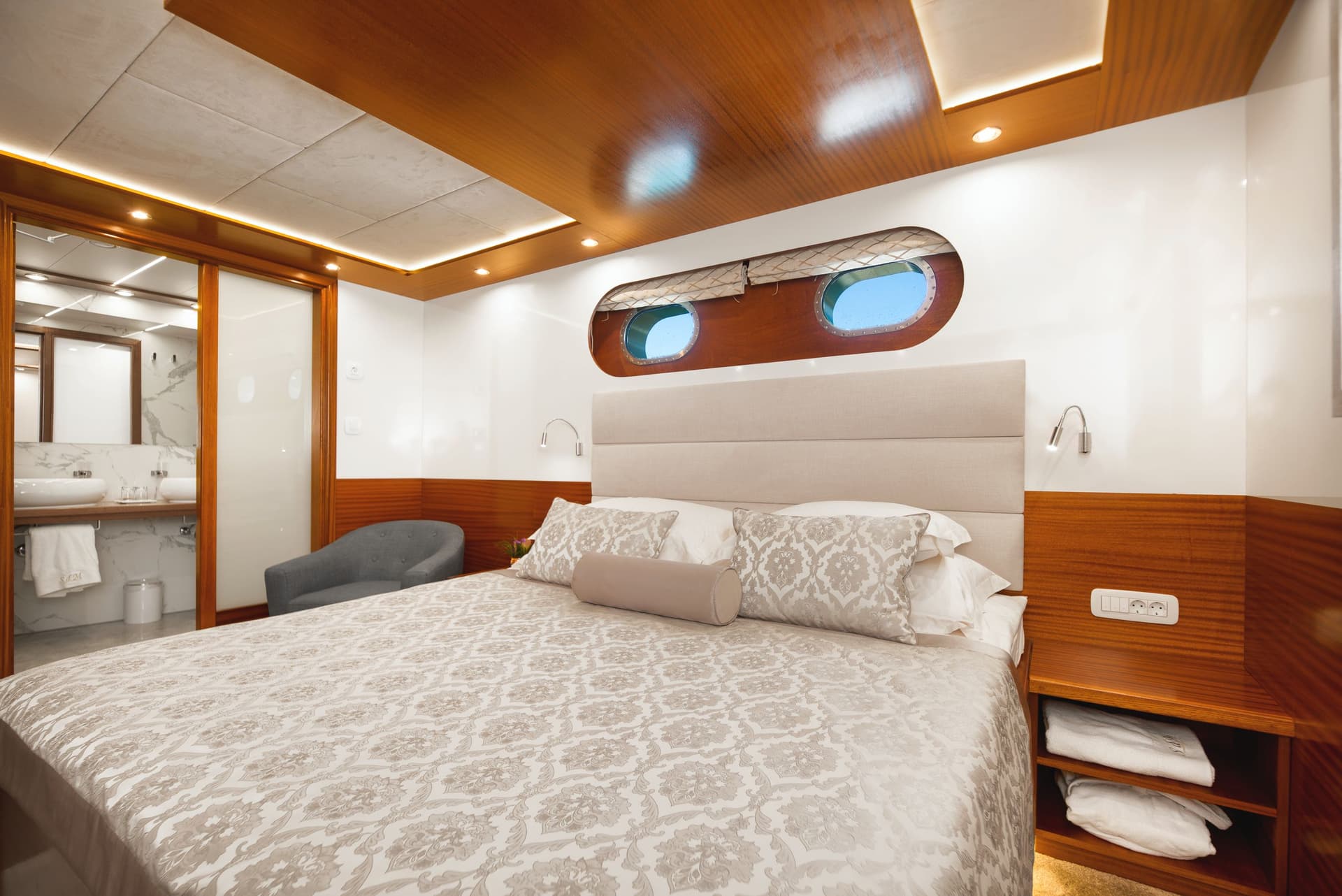Cabin bedroom with double bed, wood trim, and oval porthole windows showing blue sky.