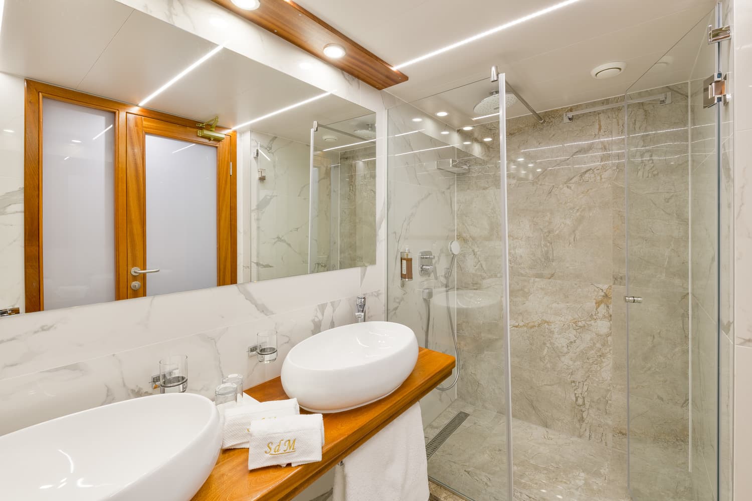 Modern bathroom with white marble, wooden counter, vessel sinks, and glass shower enclosure