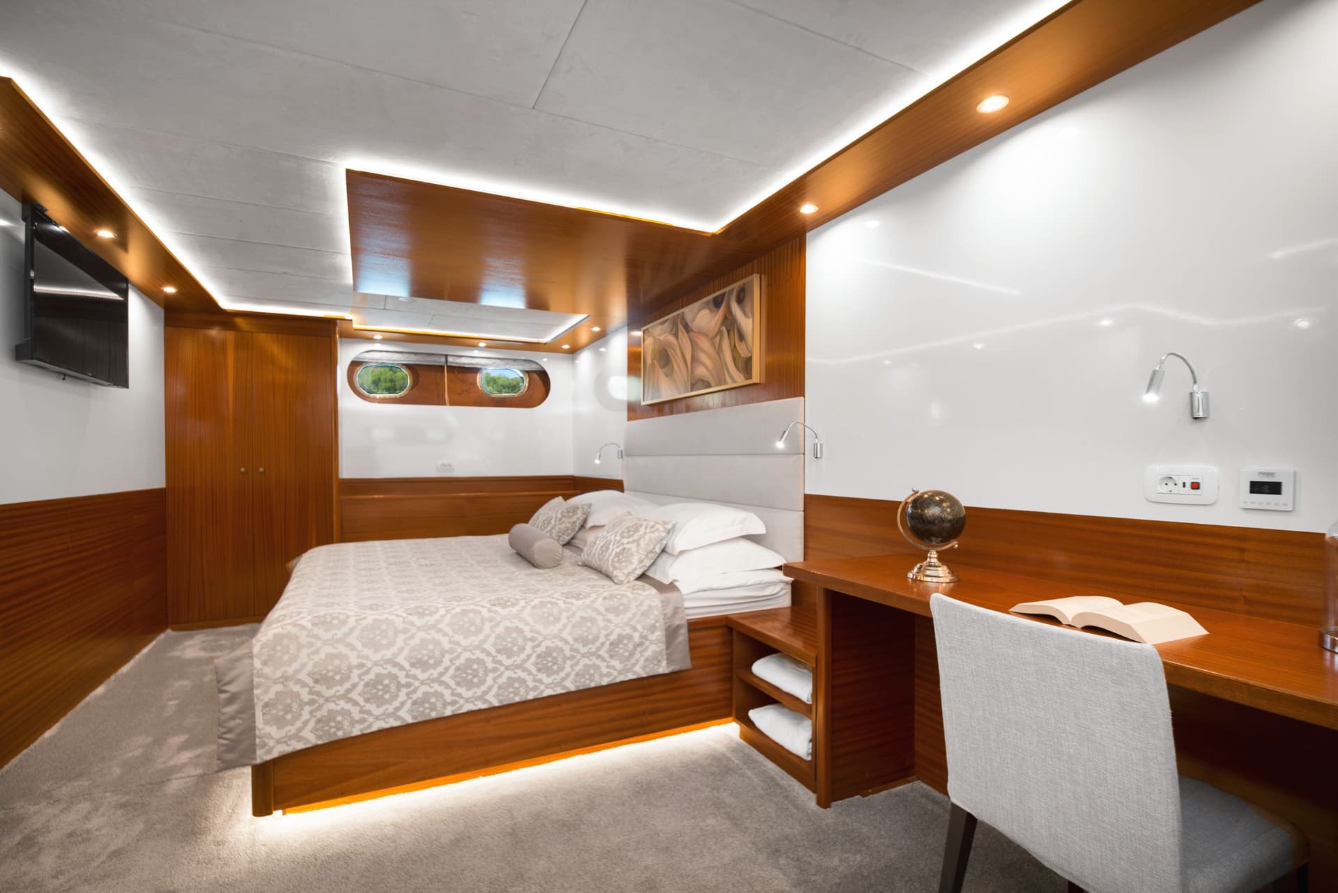 Cabin bedroom with wood paneling, plush bed, desk, and porthole windows showing greenery.