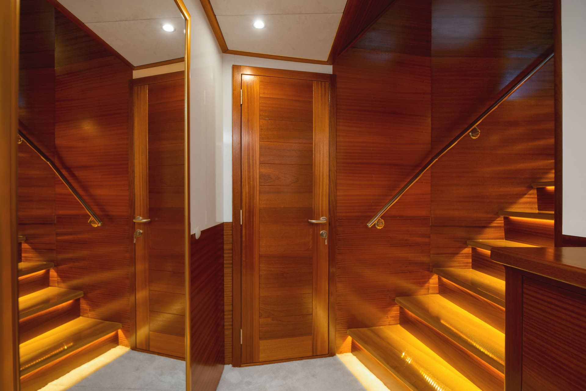 Stairway with illuminated wooden steps, polished wood paneling, and mirrored wall inside a vessel.