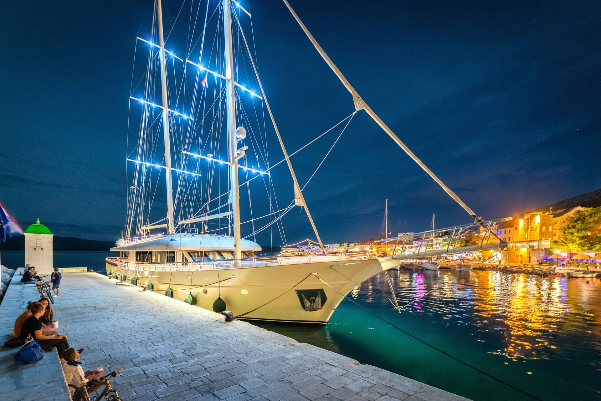 Large illuminated sailboat docked at night with people relaxing on the stone quay.