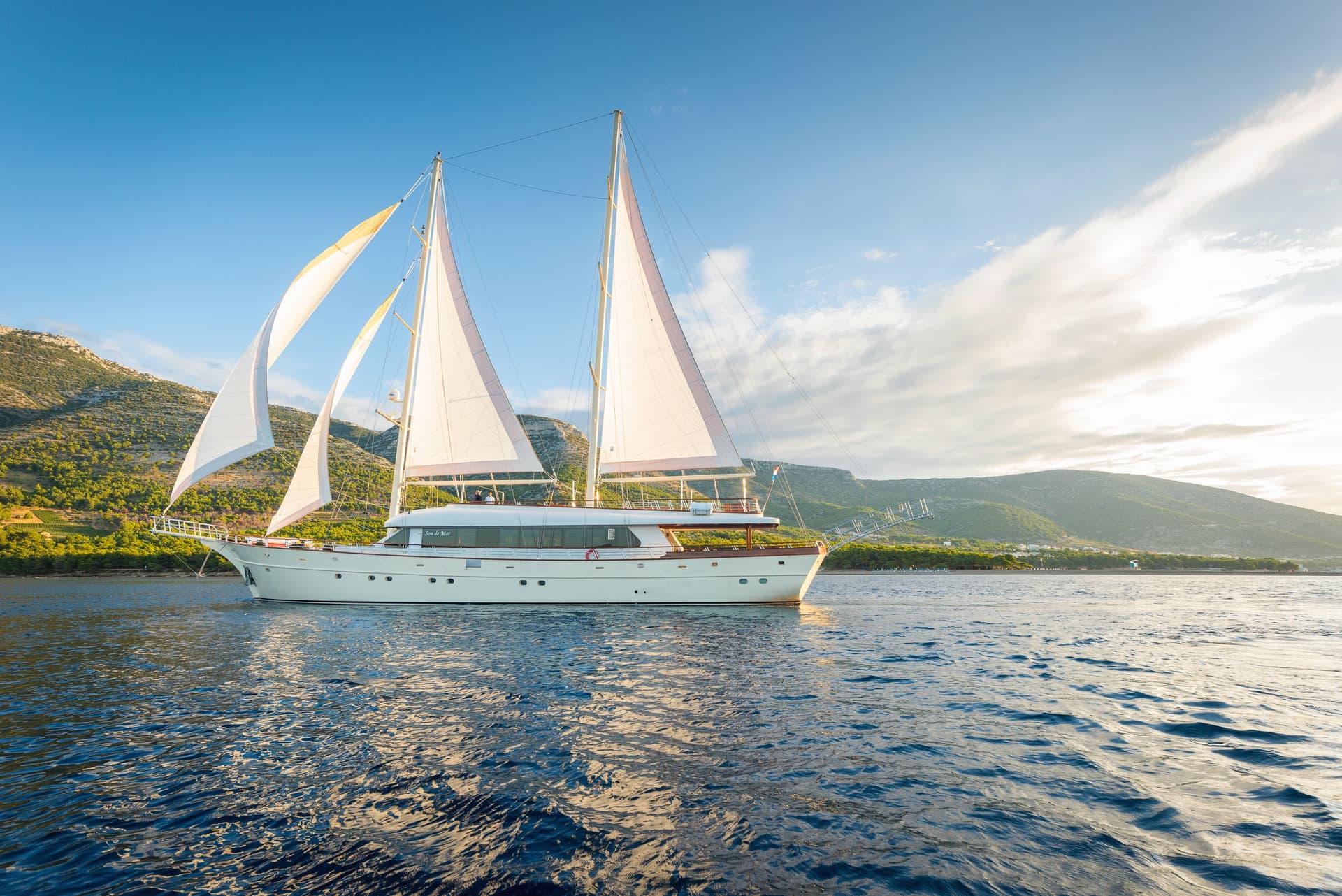 Sailboat "Son de Mar" sailing on deep blue water near a green, hilly coastline.