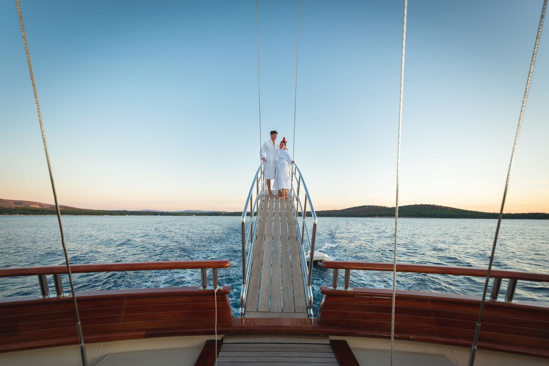 Couple in white robes on wooden swim platform of boat sailing near tree-lined coast.