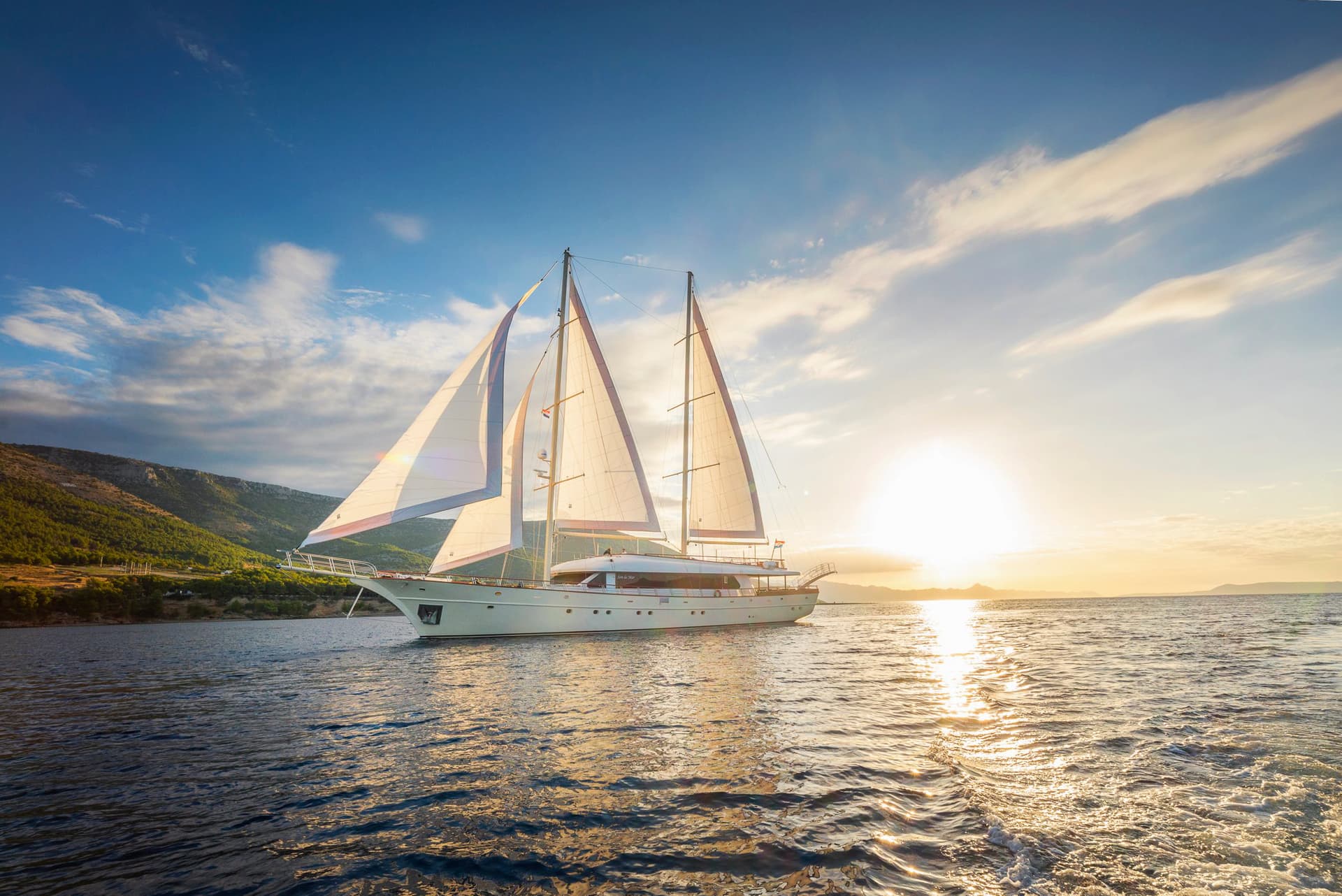 Large sailboat with sails up cruising on the sea near a green, hilly coastline at sunset in Croatia.