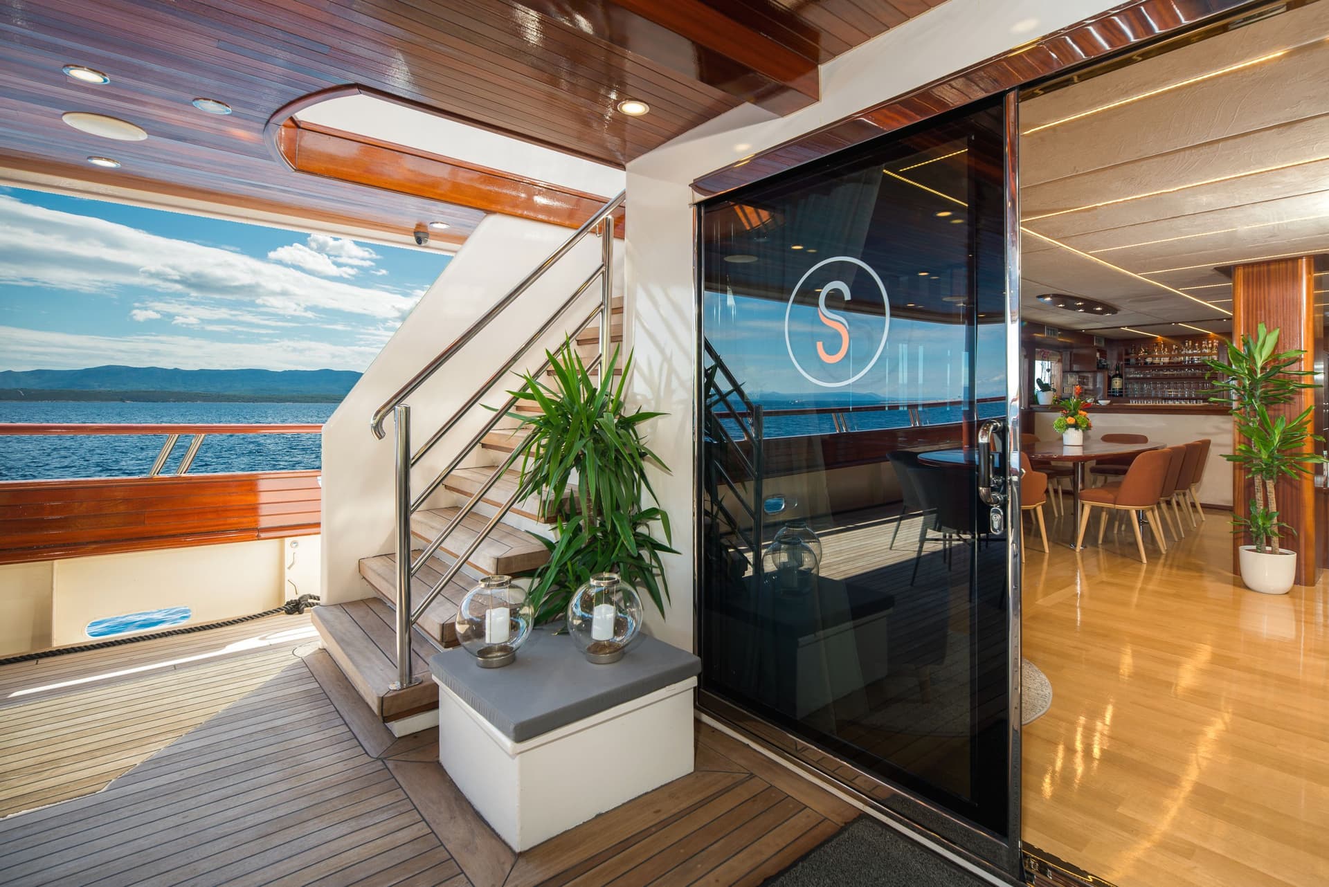 Yacht deck with teak stairs, potted plant, and view of blue sea and distant mountains.