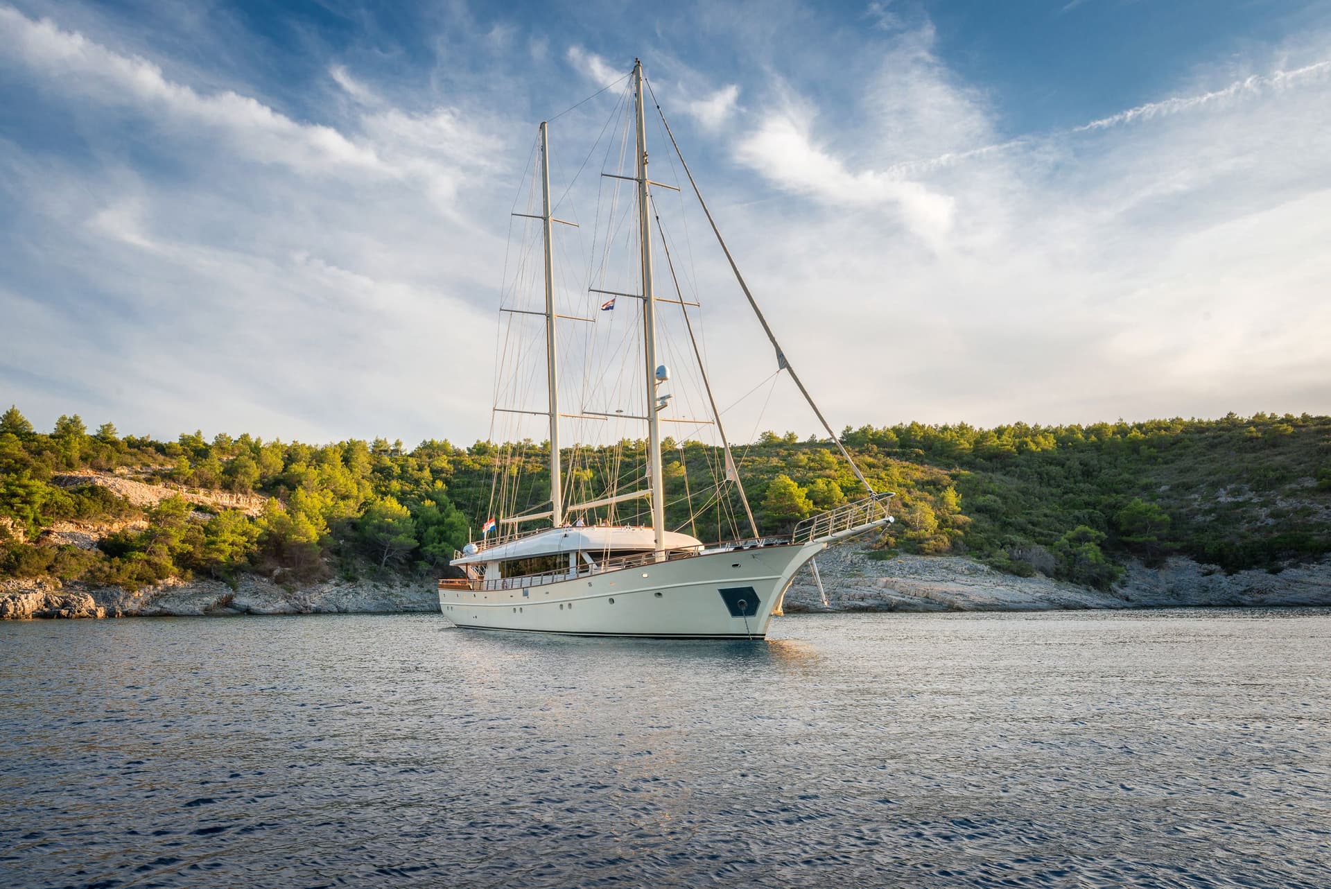 Large sailboat anchored in calm bay near rocky, forested coastline under blue sky.